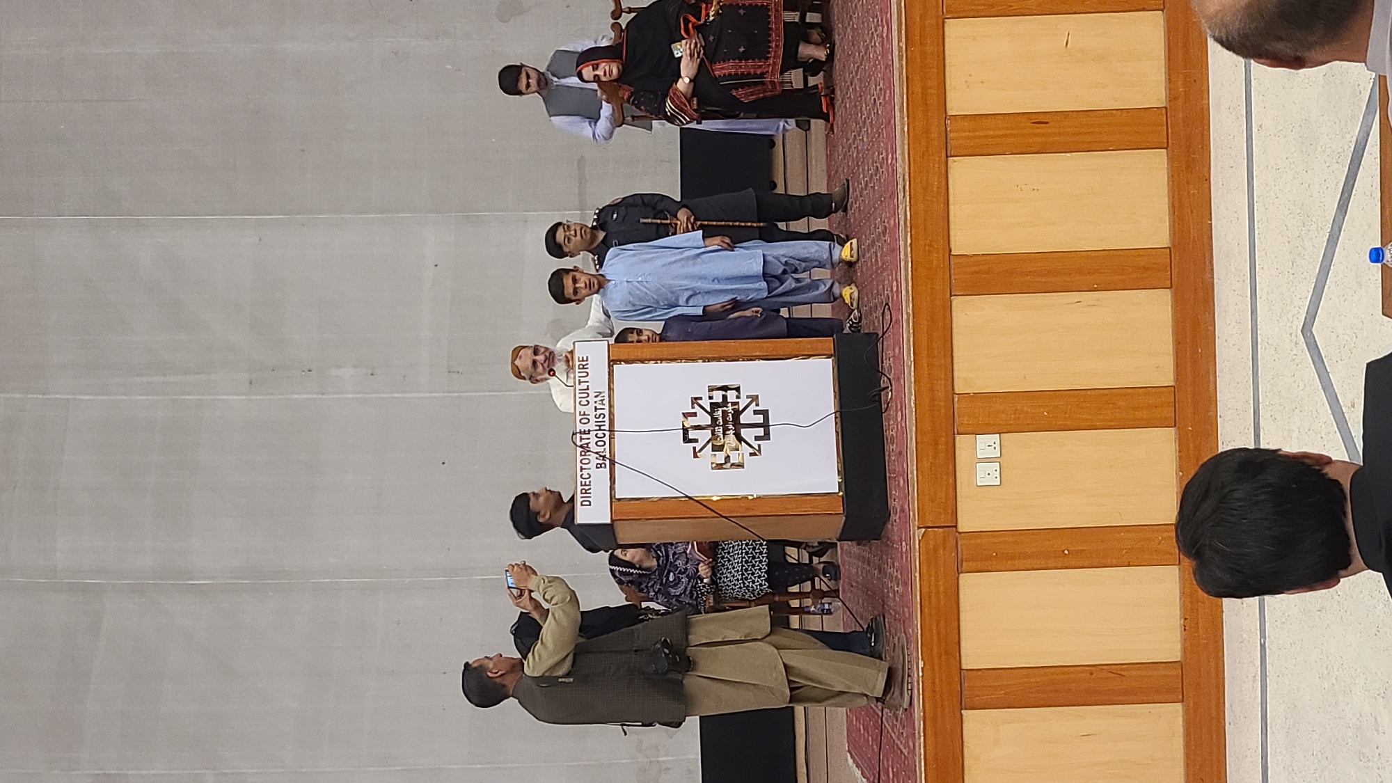 The image shows a gathering of people in Quetta City Tehsil, Pakistan. Several individuals are on a raised platform, with one man standing behind a podium that reads "Directorate of Culture Balochistan." The podium has a crest or symbol on its front. Attendees are positioned both on and off the stage. One person in the foreground is taking a picture with a phone. The surroundings include wooden paneling, outlets on the wall, and carpeting on the stage.