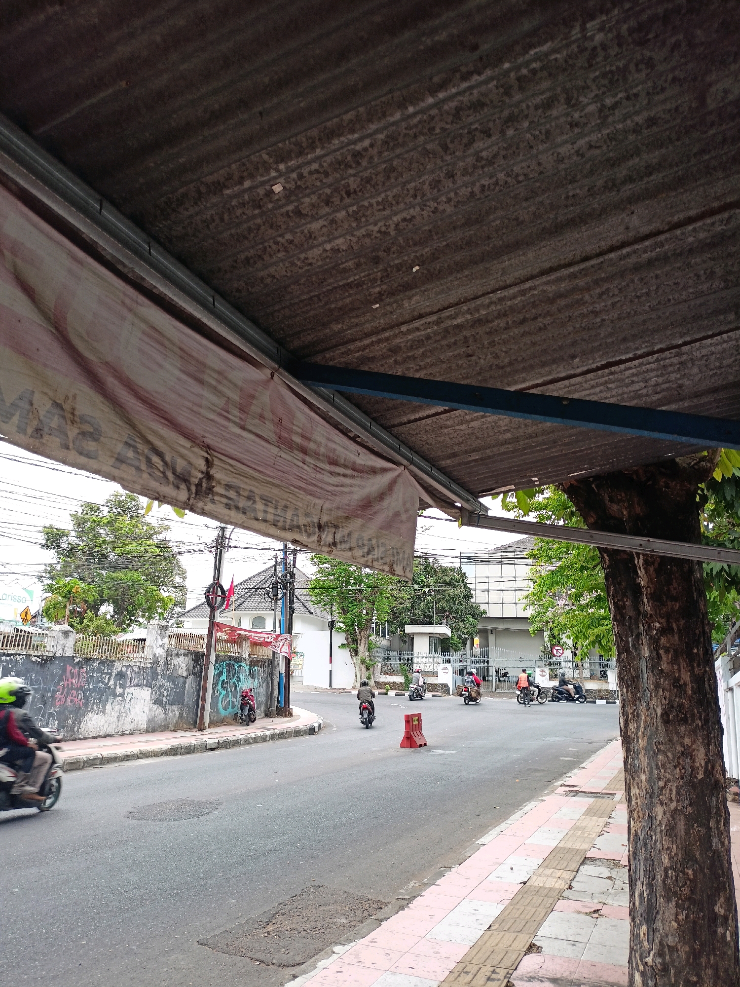 The image depicts a street scene in Magelang, Indonesia, likely taken from a covered area, possibly a bus stop or awning, indicated by the corrugated metal roof and a tattered banner visible in the foreground. The road surface is asphalt with some patchy repairs. To the left of the road, there is a sidewalk with a pink and white tiled pattern and tactile paving. Adjacent to the sidewalk is a concrete wall covered in graffiti.
Several motorcycles are in motion on the road, suggesting typical traffic flow. One motorcycle rider with a yellow helmet is visible in the left foreground, blurred due to motion. Further down the road, other motorcycles are seen, with riders and passengers. There's a red traffic cone on the road near the intersection.
In the background, buildings with modern architecture, including a light-colored building with many windows, are visible. Trees and utility poles with wires are also present, contributing to the urban environment. The overall impression is of a moderately busy street on an overcast day.