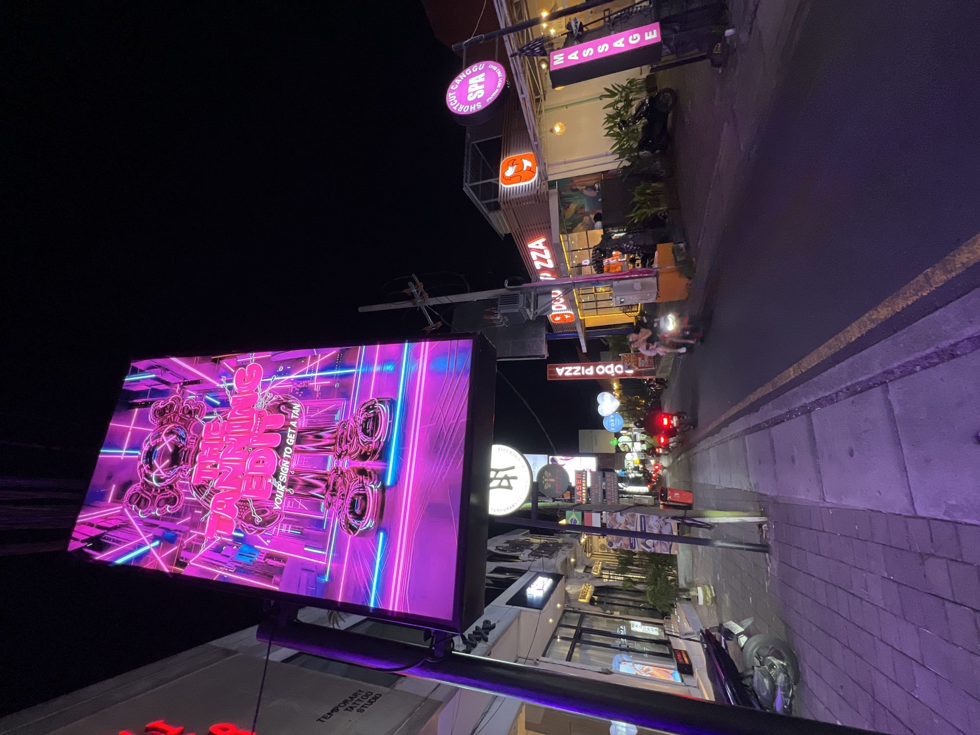 The image is taken at night in North Kuta, Indonesia. The street is lined with businesses including restaurants (Odo Pizza), spas (Canggu Shortcut Spa & Massage) and a temporary tattoo studio. A large illuminated advertising display shows a neon-themed design promoting tanning. Pedestrians and motorbikes are visible on the street. The street surface is paved and a parked motorbike is on the right.