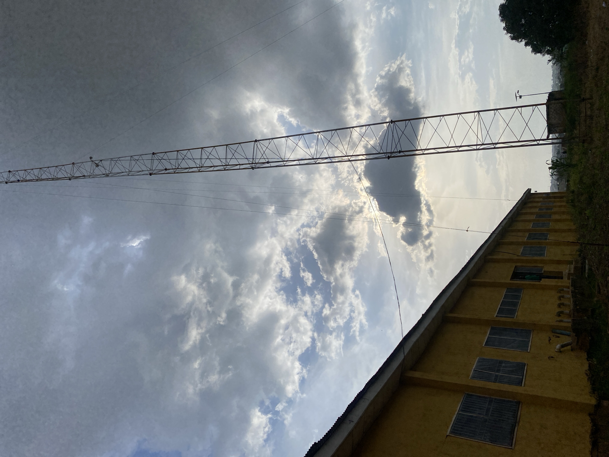 The image shows a tall, lattice tower structure against a cloudy sky. To the left, a yellow building with multiple windows runs along the lower edge of the frame. The tower is a prominent element, situated slightly to the right of the building. The sky is filled with a mix of dark and light clouds, with some sunlight breaking through. The ground is covered in grass and some vegetation. The scene appears to be outdoors, in an area that may be rural or on the outskirts of a town. While the location is identified as Rafin Par, Nigeria, there are no specific landmarks or discernible features within the image that directly confirm this exact location. There are no visible people or interactions occurring in the scene.