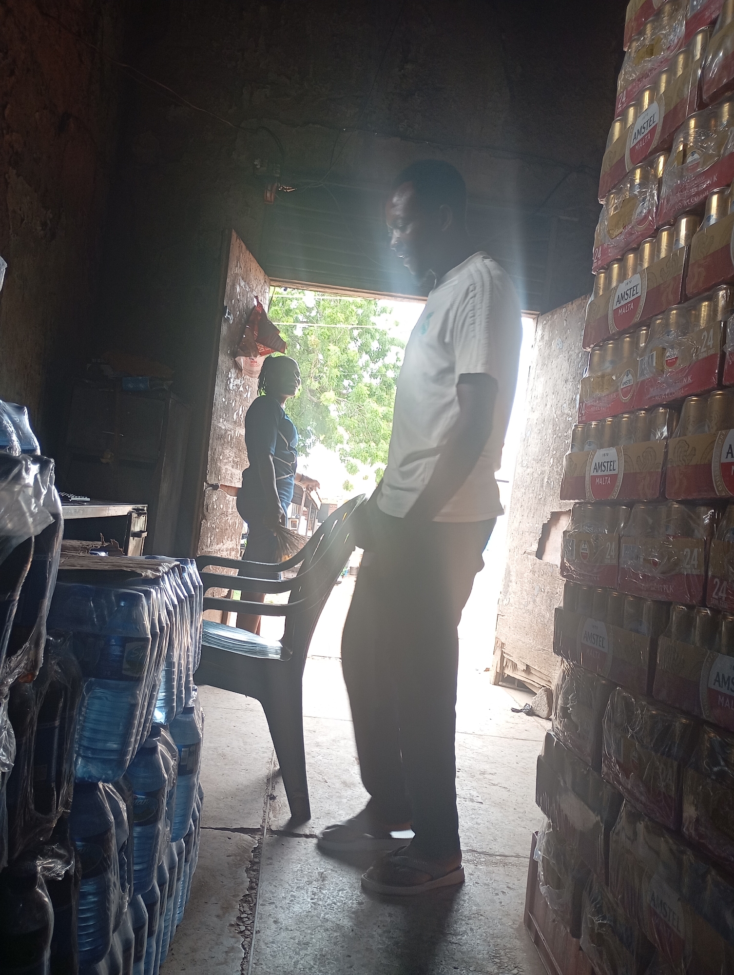 A man and a woman are standing inside a dimly lit warehouse, which appears to be a storage facility for beverages. The man, wearing a white t-shirt and dark pants, is looking towards the right side of the image. The woman, wearing a dark t-shirt and blue shorts, is standing near a chair and holding a broom. Stacked high on the right side of the warehouse are numerous cases of Amstel Malta, a malt beverage, wrapped in plastic. On the left side of the warehouse, stacks of bottled water are also visible, also wrapped in plastic. The environment is rustic, with concrete floors and exposed walls. The location is identified as Shendam, Nigeria.