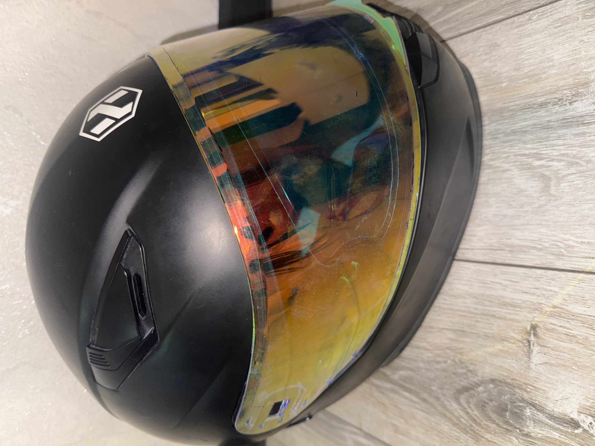 A black motorcycle helmet with a reflective yellow-orange visor sits on a wooden floor. The helmet has a white logo on the right side, which appears to be an octagon with an abstract "X" design inside. There are some scuff marks and smudges visible on the visor. The background is a textured, light-colored wall. The image appears to be a close-up shot, taken indoors, likely in a residential setting. There are no people or animals present in the image. The location is not identifiable as Bishkek City, Kyrgyzstan, as there are no identifying landmarks or contextual clues.