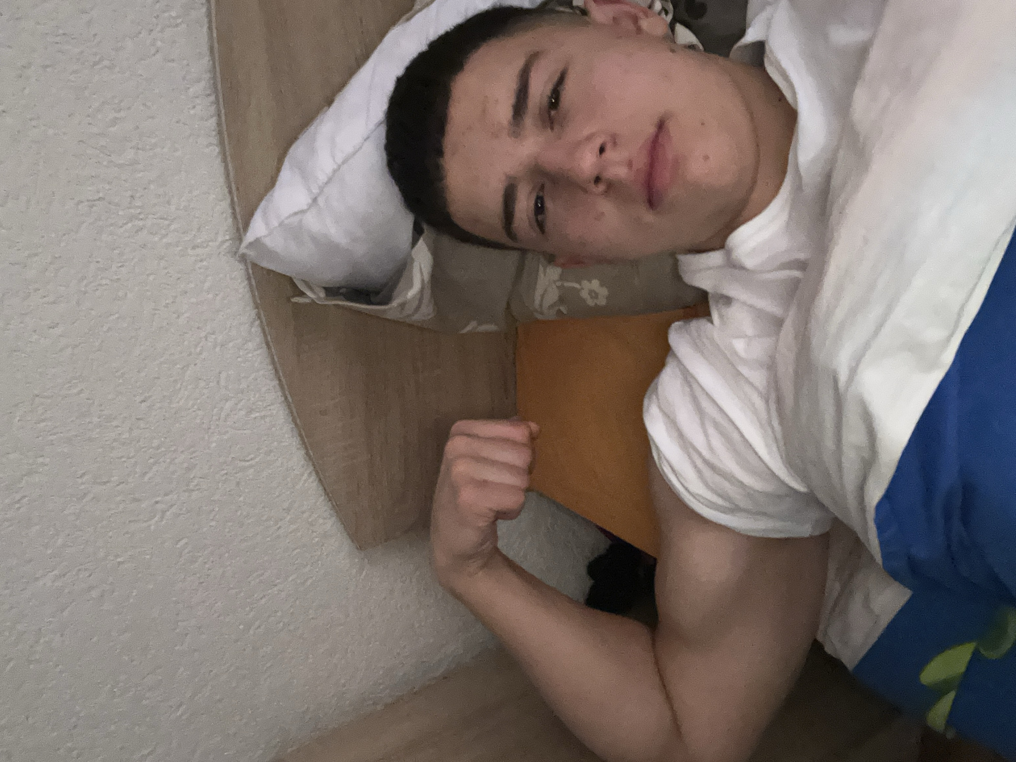 A young man with a close-cropped dark haircut is lying in bed. He is wearing a white t-shirt and is partially covered by a blue blanket. His right arm is flexed, displaying his bicep. The bed has a white pillow, an orange cushion, and a headboard. The background shows a beige wall. There is no information to determine that the image was taken in Zlatograd, Bulgaria.
