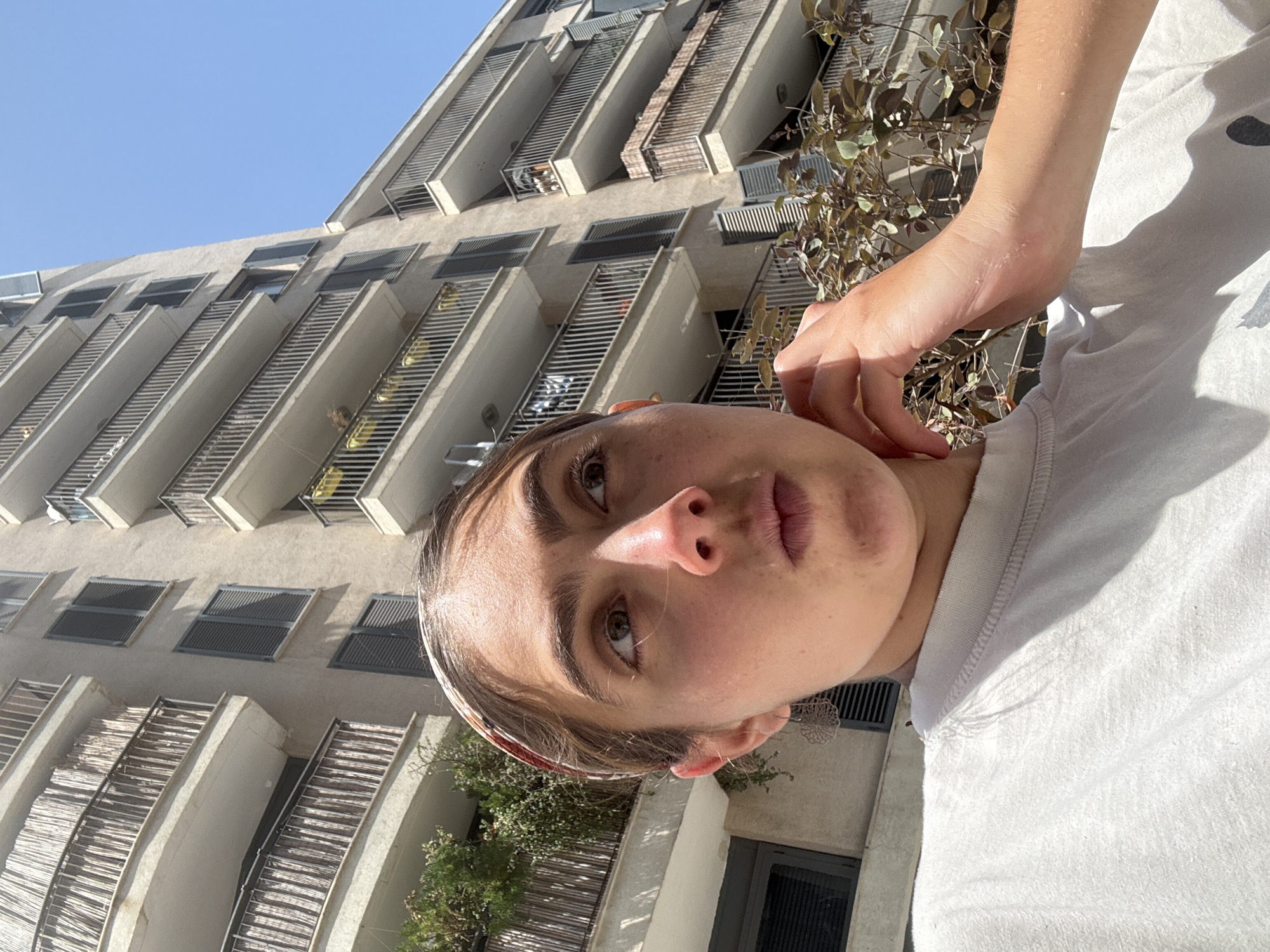 A close-up, angled shot shows a person, likely female, in front of a tall apartment building. The person has fair skin, light brown hair with a patterned headband, and appears to be wearing a white t-shirt. The building is light-colored with numerous balconies featuring slatted railings. Some balconies have solar panels or privacy screens. Foliage is visible on some of the balconies. The sky is clear and blue. The image's angle suggests it was taken from a low perspective. The architectural style and environmental elements suggest the location is likely in Tel Aviv, Israel.