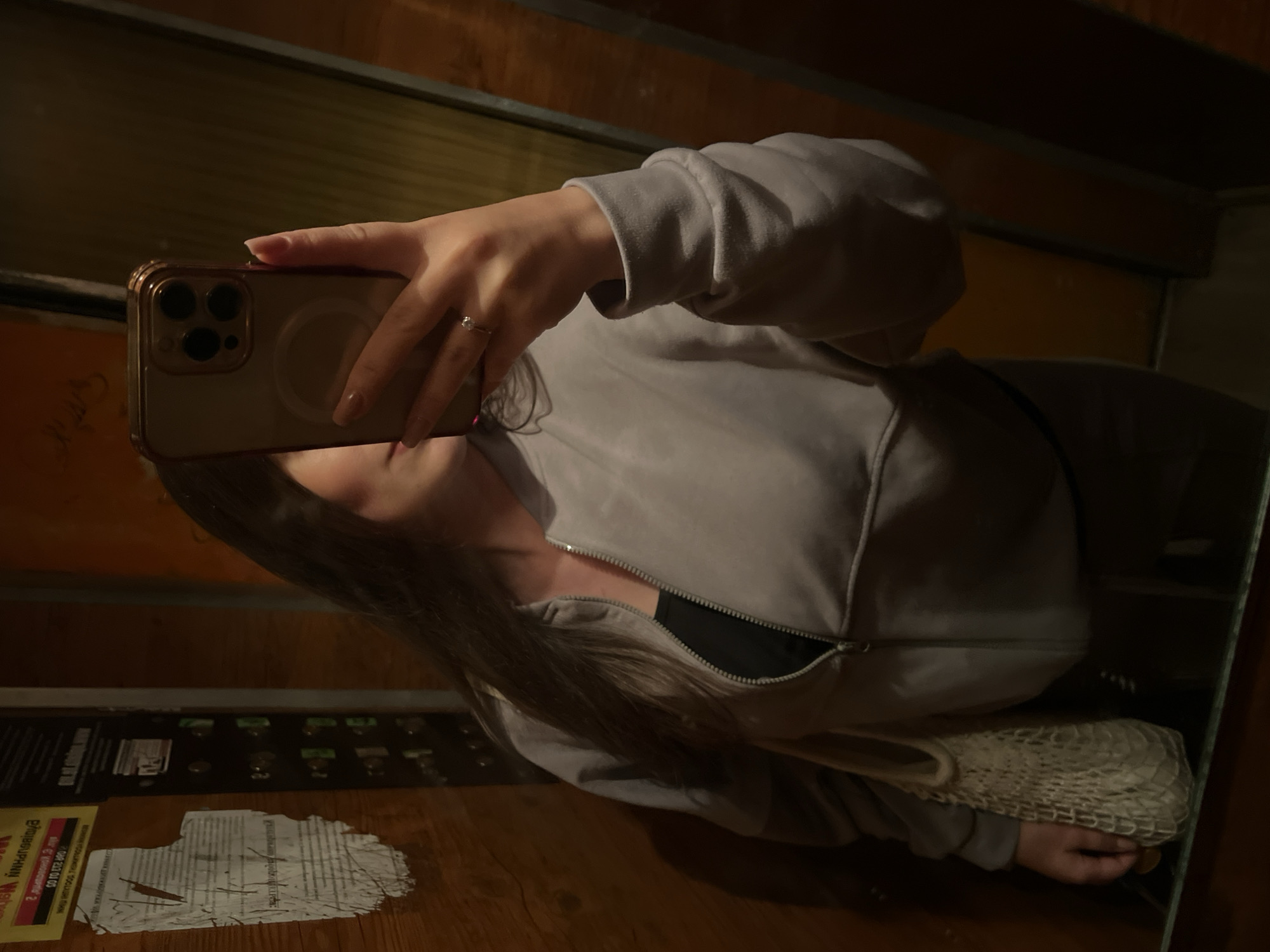 A person is taking a mirror selfie in an elevator with wooden paneling and what appears to be a notice board on the left side. The person is wearing a light grey zip-up hoodie over a black top and is holding a smartphone in their right hand. A ring is visible on their ring finger. A mesh bag is slung over their left shoulder. The elevator door frame is visible on the right. The scene appears to be indoors, likely in an urban setting. The notice board on the left has some printed text and is partially torn. Based on the distinct elevator paneling and the style of the notice board, this elevator is located in a residential building in Ternopil, Ukraine.