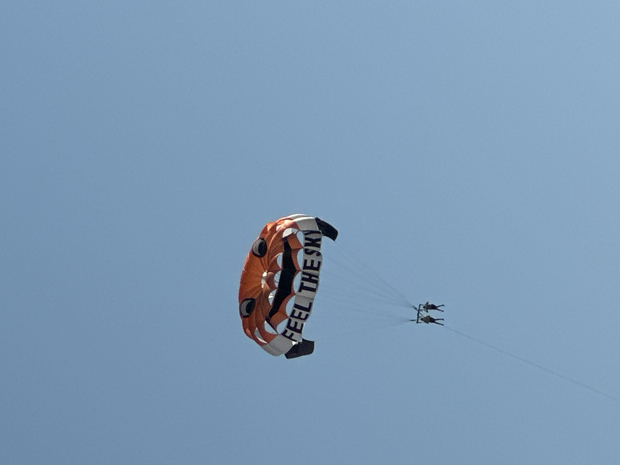 A parasail with a smiling face design and the words "FEEL THE SKY" is airborne against a clear blue sky. Two individuals are suspended beneath the parasail. The image appears to be taken from a low angle, looking up at the parasailing activity. The location is not discernible from the image alone.