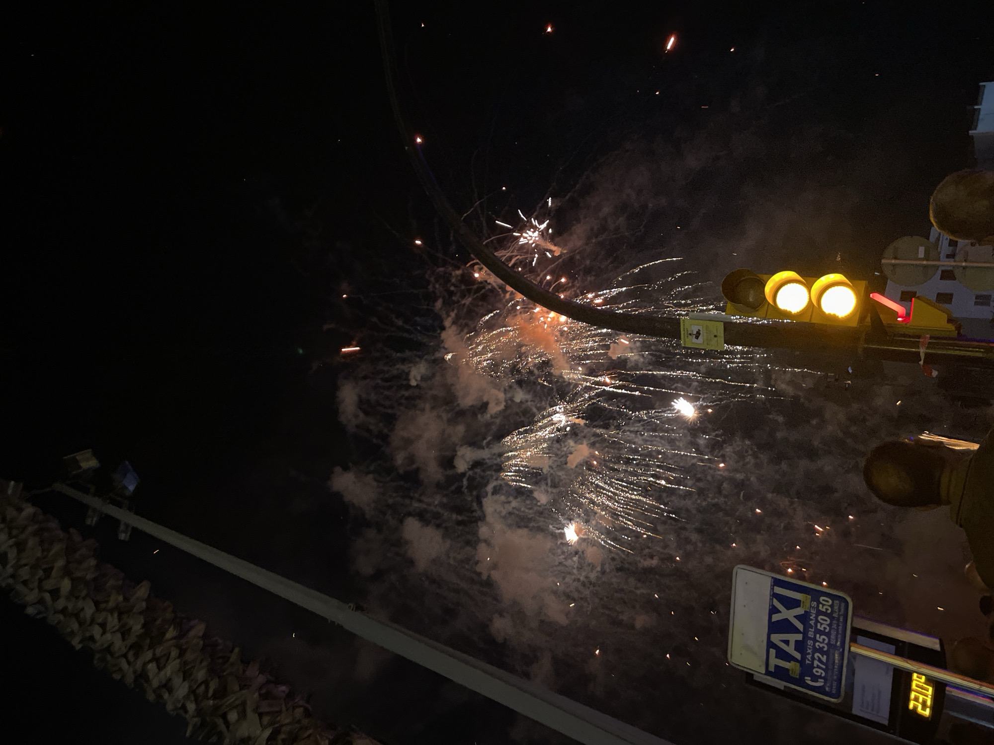 A night time scene captures fireworks exploding in the dark sky over Blanes, Spain. A palm tree frond is visible in the upper left corner. A tall street light pole with two light fixtures stands prominently on the left side of the image, angled towards the right. A traffic light, with its two yellow lights illuminated, is positioned centrally and slightly to the right. In the lower left quadrant, a taxi sign with "TAXI" in large blue letters and a phone number is mounted on a pole, accompanied by a digital display showing "23.0°". Several people are visible in the lower part of the image, with their backs to the camera, looking up at the fireworks. The fireworks themselves are a mix of bright white and orange trails and bursts, with smoke clouds diffusing the light. The overall environment suggests an outdoor event or celebration.