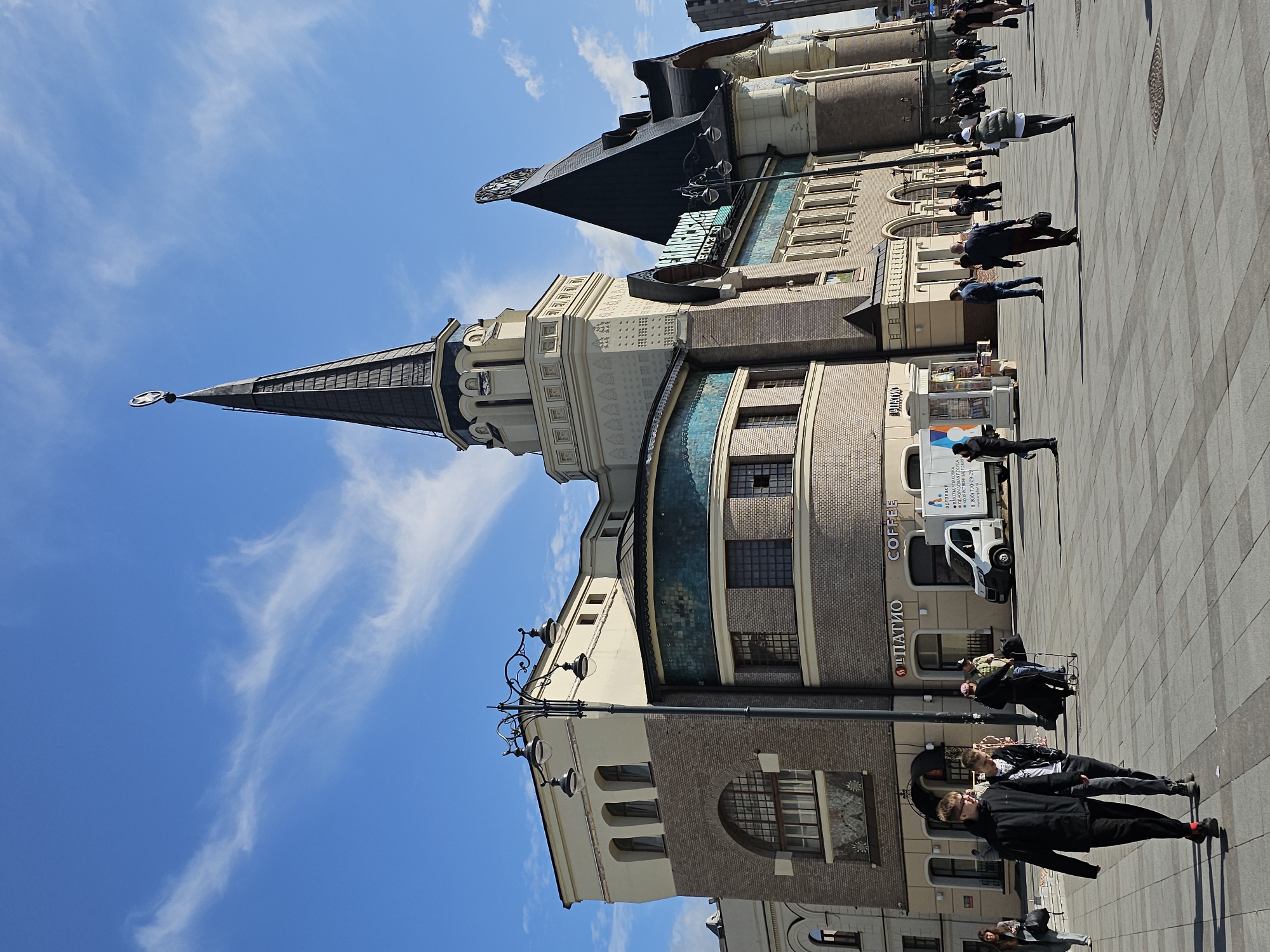 This is a wide-angle, eye-level, outdoor shot of the Kazansky railway station in Moscow, Russia. The station, with its distinctive spired tower and patterned facade, dominates the frame under a bright blue sky with scattered white clouds. A large paved plaza is in the foreground, with several people walking or standing. On the left side of the plaza, two young men in dark clothing are walking towards the camera, with one gesturing with his left hand. To their right, a white delivery van with a "Coffee" sign is parked near a small kiosk. Other pedestrians are scattered across the plaza, some walking away from the station and others towards it. A tall, ornate lamppost stands to the left of the station's entrance. The architecture of the station is detailed, featuring brickwork, arched windows, and decorative tiling. The overall scene depicts a busy urban environment on a clear day.