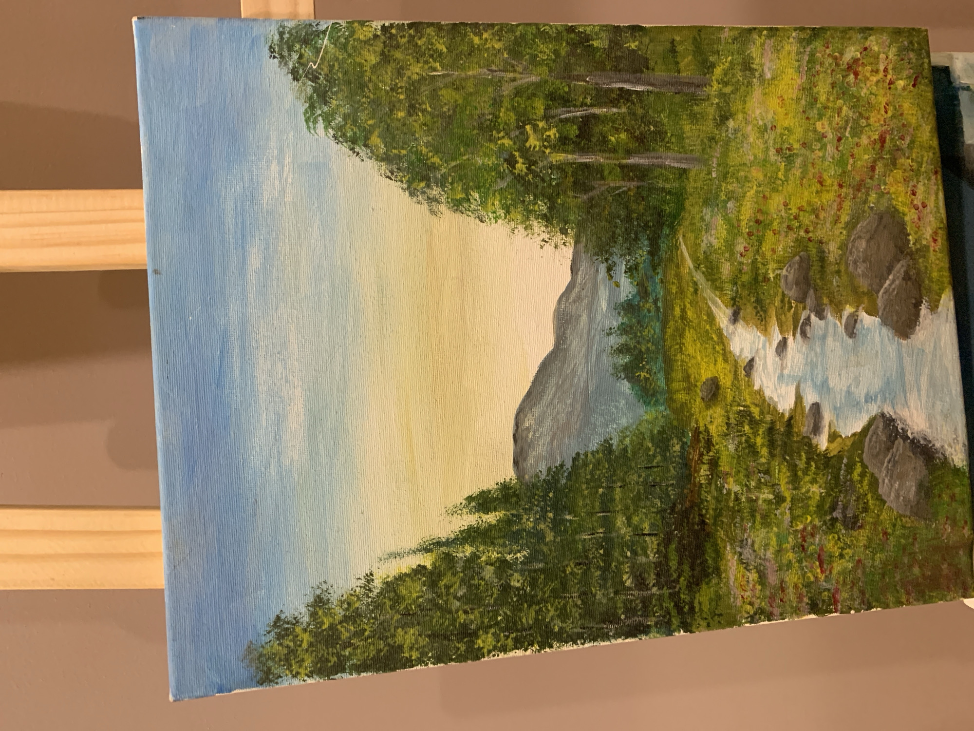 A painting on canvas depicts a serene natural landscape. The scene is dominated by a winding stream flowing through a meadow dotted with wildflowers and rocks. Tall green trees frame the sides of the composition, with a range of mountains visible in the distance under a gradient sky that transitions from blue at the top to a soft yellow near the horizon. The painting is displayed on a wooden easel, with the background appearing as a plain, light brown wall. The location of Aketu-Oja, Nigeria is not depicted in this image.