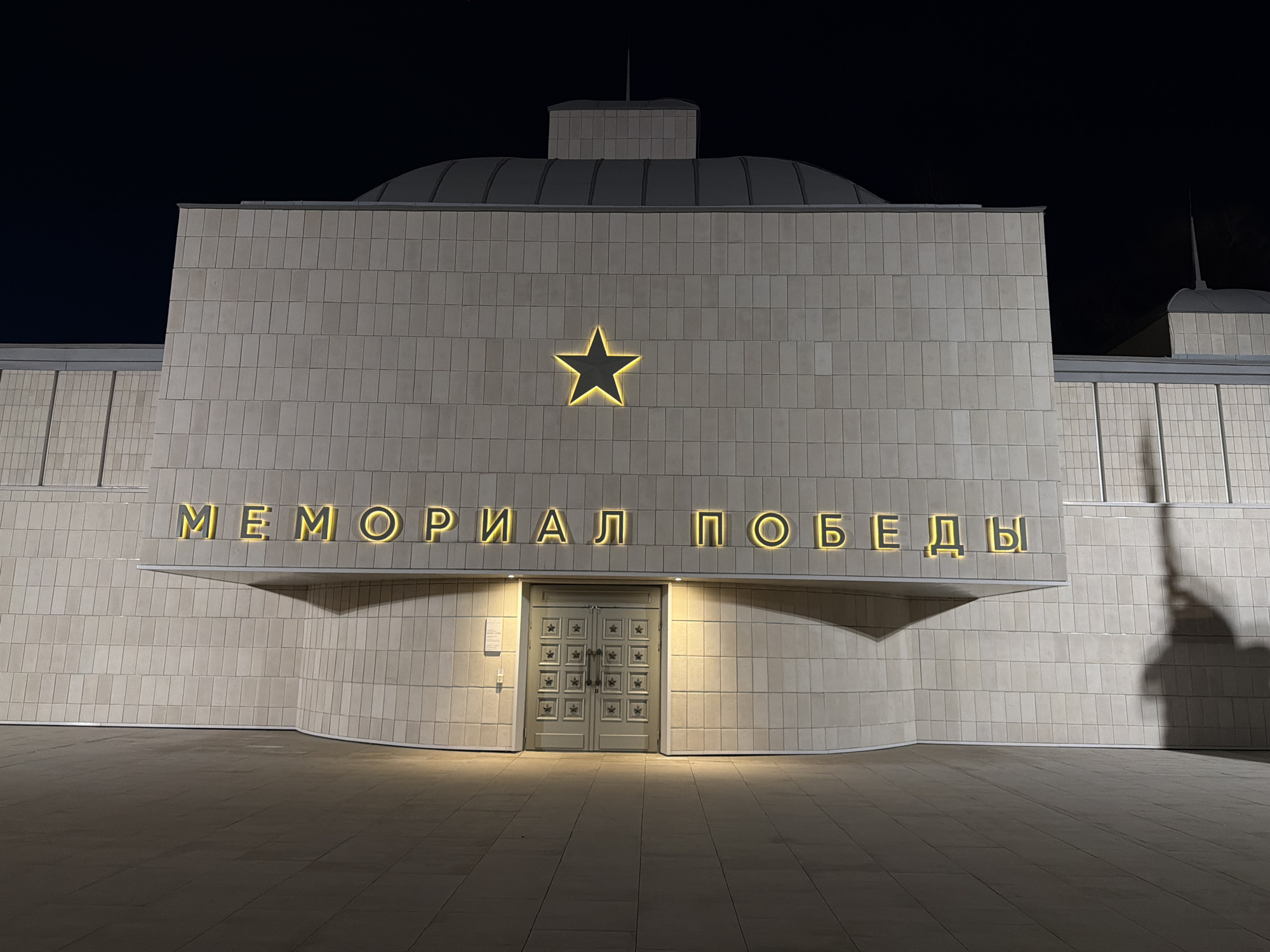 A low-angle, wide shot shows the front entrance of the Memorial Pobedy (Victory Memorial) in Krasnoyarsk, Russia, at night. The building is a pale greyish-white stone or tile, with a large, lighted five-point star centered on the main facade above the double doors. Above the doors, the words "МЕМОРИАЛ ПОБЕДЫ" are displayed in lighted, gold-colored Cyrillic letters. The building has a domed roof behind the main facade. The building and entrance area are well-lit, and the ground is a dark grey color. A shadow appears on the right side of the building. The sky is dark, and no people are visible.