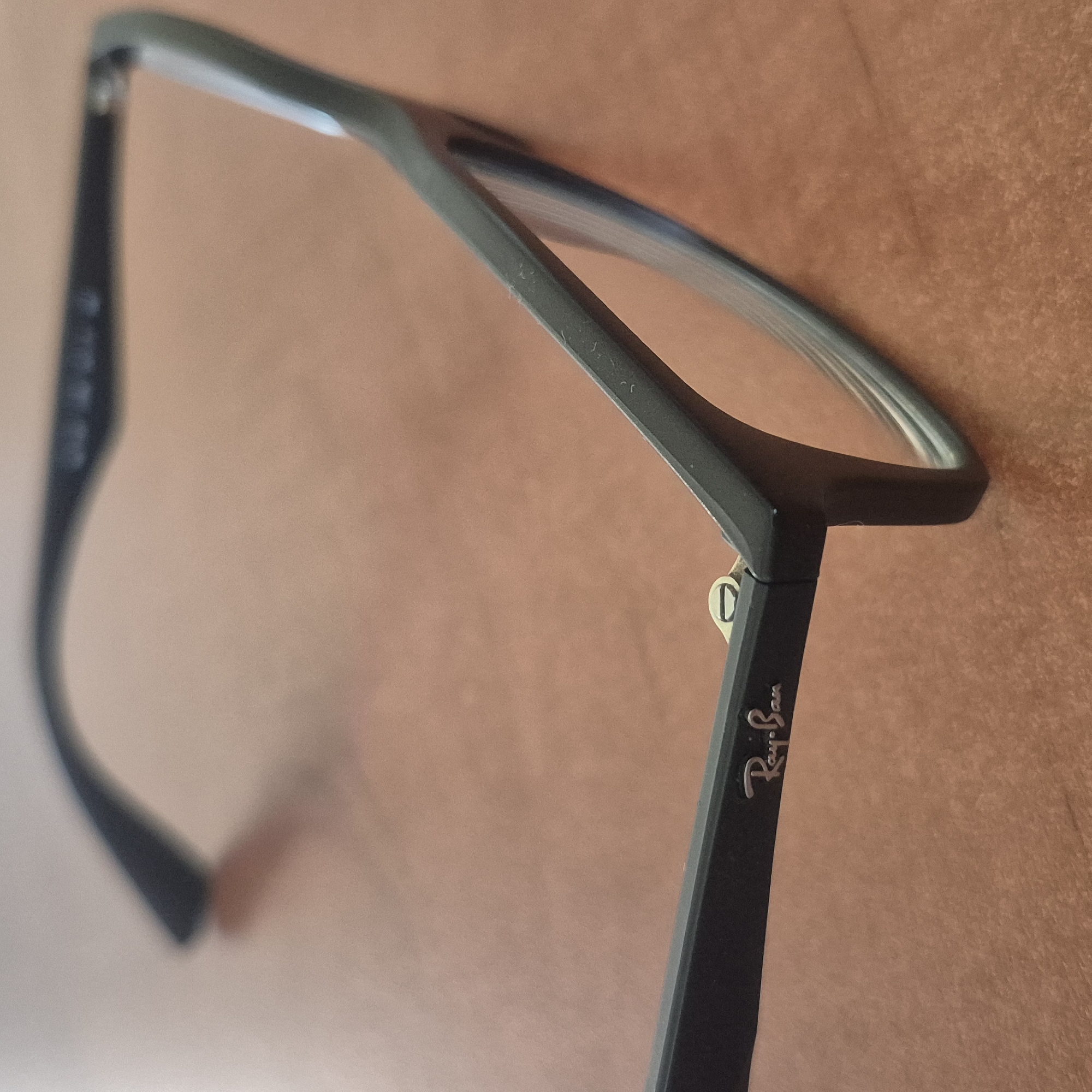 A close-up, angled shot shows a pair of black Ray-Ban eyeglasses resting on a textured, reddish-brown surface that resembles wood or cork. The eyeglasses are dark and appear to be made of plastic. The left temple of the glasses is partially visible, showing some faint markings. The right temple is more prominent, displaying the iconic silver Ray-Ban logo in script, with the words "Ray-Ban" clearly legible. A small, metallic hinge mechanism is also visible where the temple connects to the frame. The lenses are clear and reflect some light, indicating they are likely prescription lenses. The overall lighting is soft, suggesting an indoor setting, possibly a desk or table. There are no people visible in the frame. The context provided, Nowa Wieś Książęca, Poland, is a factual piece of information associated with the image, but it does not offer visual cues within the image itself.