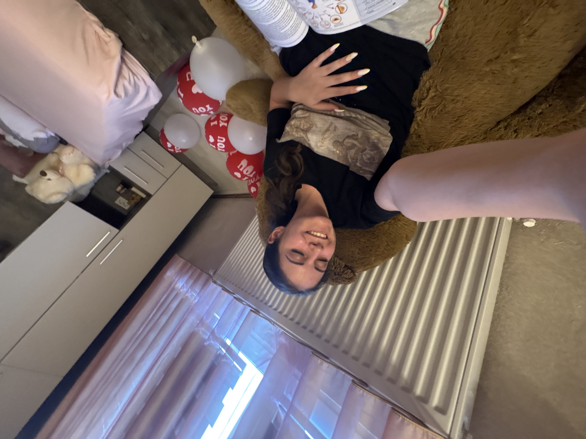 The image shows a young woman lying down on a large, brown, furry object, possibly a stuffed bear. She is wearing a black t-shirt with a printed design and grey shorts with a striped waistband. Her eyes are closed, and she is smiling. Scattered around her are several red and white balloons, some of which are printed with "I LOVE YOU." In the background, a bed with pink bedding and a large stuffed white bear are visible. A dresser with drawers and a window with pink curtains are also in the scene. The overall setting appears to be a bedroom. This photograph was taken in Bulgarovo, Bulgaria.