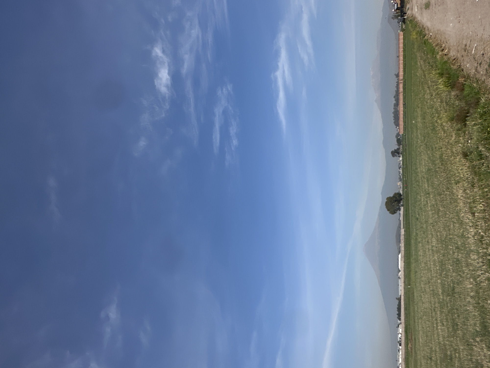 This media file captures a wide, open landscape under a bright, clear blue sky. The sky, occupying the majority of the frame, features scattered, wispy white clouds that are thin and streaky, characteristic of cirrus or altocumulus formations. The sky's color graduates from a deeper blue in the upper left to a paler, more washed-out hue towards the horizon, suggesting atmospheric haze.
In the foreground, a vast, flat, green field stretches across the bottom portion of the image. The field appears to be a cultivated area, possibly grassland or crops, and is bordered on the right by a dry, lighter-colored dirt path or road, which separates it from a patch of wilder grass and earth.
The background is dominated by a range of distant mountains, which appear muted blue-grey and hazy due to atmospheric perspective. A distinct, somewhat conical peak is visible among these mountains towards the center-right. At the foothills of the mountains, faint, indistinct structures and a few scattered trees can be seen, hinting at a distant settlement or rural development. One more prominent, rounded tree stands out in the mid-ground against the mountain backdrop.
The scene depicts a peaceful daytime setting with clear weather, although atmospheric haze affects visibility of distant objects. There is no visible activity or identifiable individuals. No text is visible in the image. The wide fields and distant mountains are consistent with the provided location context of San Luis Tehuiloyocan, Mexico, suggesting a view of the surrounding agricultural land and possibly volcanoes like Popocatépetl or Iztaccíhuatl, or other mountains in the region.