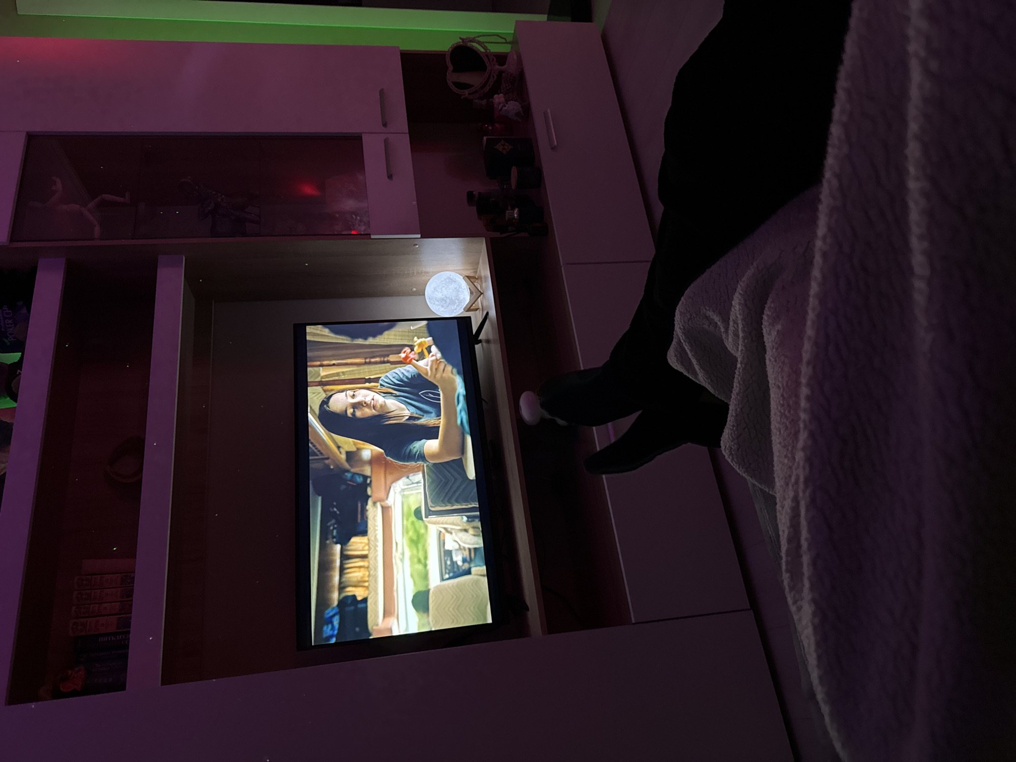 The image captures an indoor scene, likely a living room or bedroom, illuminated by artificial light in the evening or at night. A person is visible on the right side of the frame, lying on a couch or bed, mostly covered by a white, textured blanket. Their legs, clad in dark pants and socks or slippers, are visible.
A flat-screen television is centrally positioned within a light-colored, built-in shelving unit. The TV displays a woman with dark hair, wearing a casual top, looking upwards while holding a small object in her hands. The background on the screen suggests the interior of a vehicle, possibly an RV or camper van, with visible seating and windows.
To the right of the television, on a shelf, a glowing spherical lamp designed to resemble the moon provides a soft light. The shelving unit contains various items, including what appear to be decorative figurines or sculptures behind a reflective panel on the upper shelves. On a lower shelf to the left of the TV, a stack of books or DVDs is present, with partially legible text on one spine reading "PORKER CH..." and another line possibly "ИНТЕРНЕТ" in Cyrillic script.
The room is softly lit with green LED strip lighting at the very top of the wall and a purple/pink glow cast over the shelving units, creating a relaxed and cozy atmosphere.