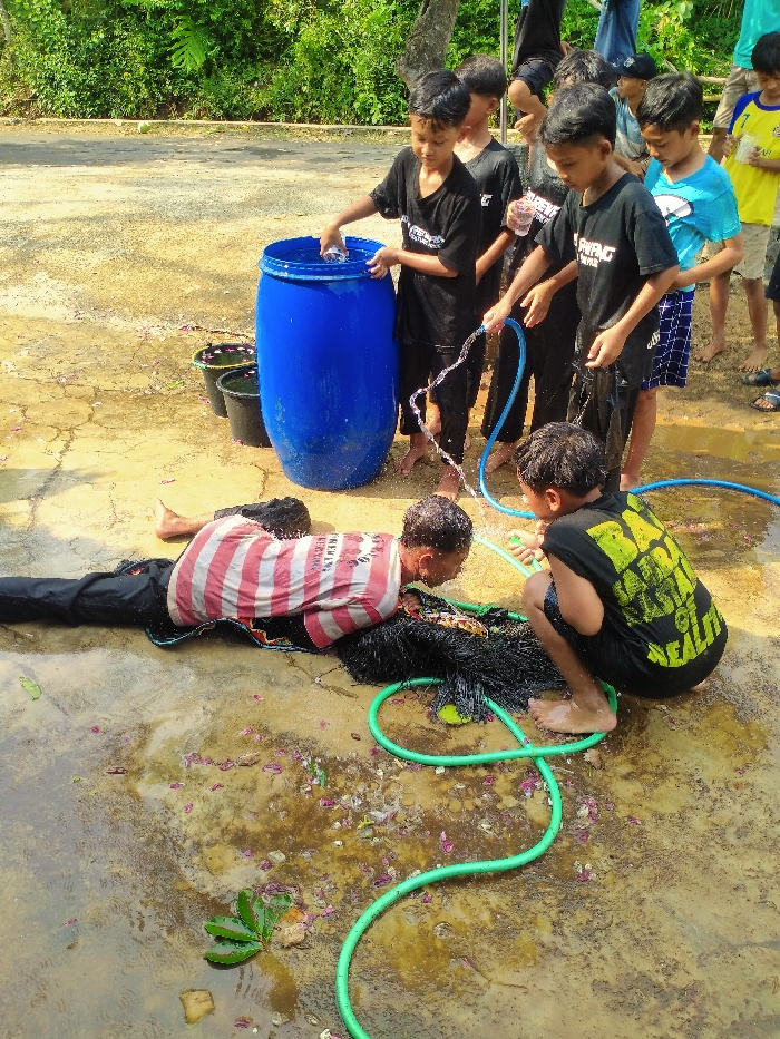 The scene, located outdoors in Gebyog, Indonesia, features at least eight young males engaged in an activity involving water. The ground is a wet, uneven paved surface, marked by brownish stains and scattered pink and white flower petals, along with green leaves.
In the foreground, one young male is lying prone, face down, wearing a red and white striped short-sleeved shirt and dark pants. His head rests on a damp, black, tangled net or fabric. Adjacent to him, a second young male, clad in a black sleeveless shirt with yellow text, is crouched, holding a green garden hose, and directing a stream of water onto the prone individual's head and upper back.
Behind them, a blue plastic barrel filled with water stands upright. A third young male, wearing a black T-shirt, is positioned next to the barrel, reaching into its opening. A fourth young male, also in a black T-shirt, holds a blue garden hose, from which water is actively spraying towards the ground. Several other young males, one in a light blue T-shirt and another in a yellow T-shirt, are visible standing in the background, observing the scene. Most participants appear wet, and some are barefoot. In the background, green foliage is visible.