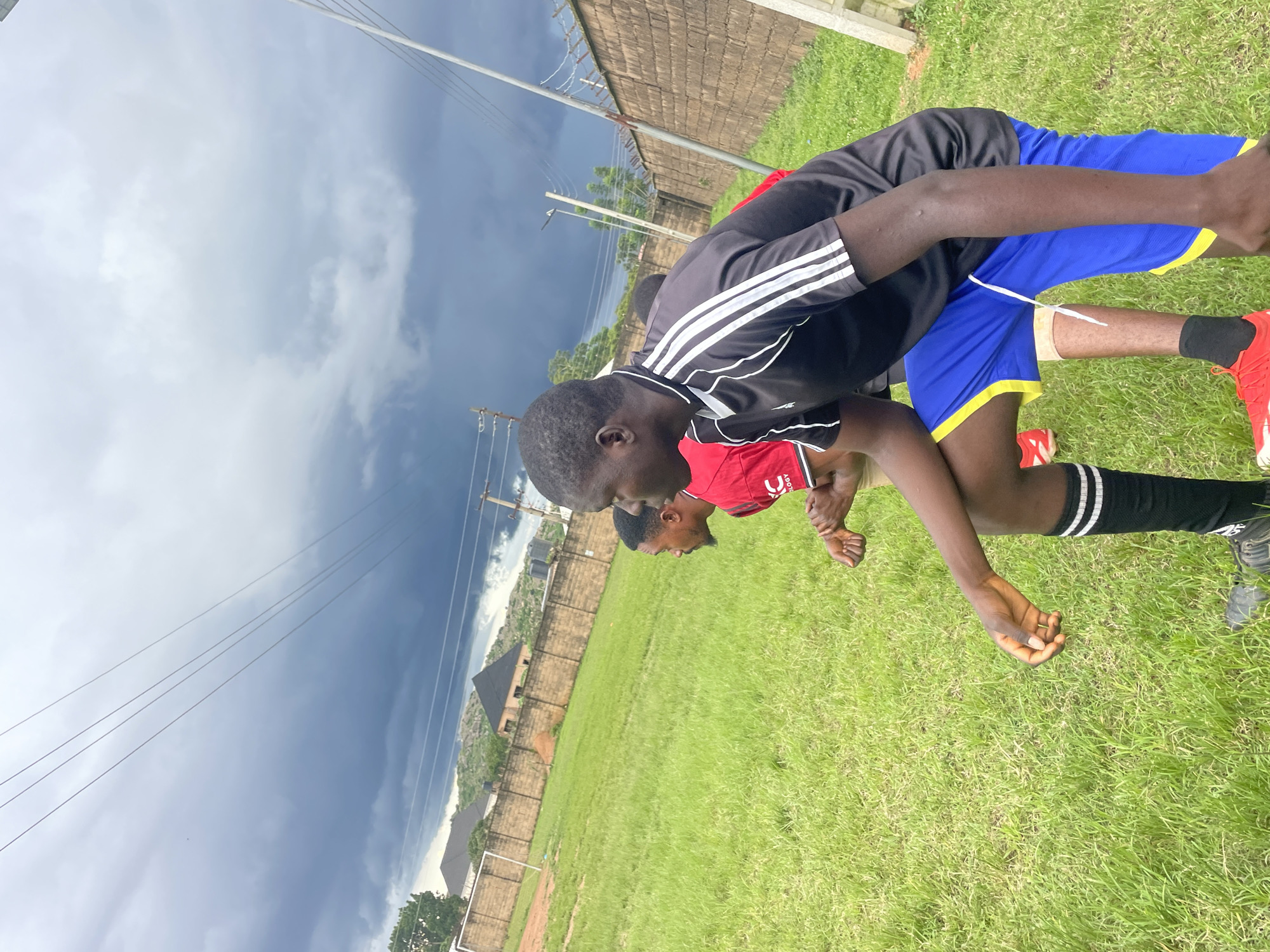 This is a full, eye-level shot of three young men on a green grass field, possibly a sports field. The sky above is dark and ominous, suggesting an approaching storm. The men are dressed in athletic wear, two in shirts and shorts, and one in a black shirt with white stripes and blue shorts. One man is in the foreground, his body turned to the right, looking off-camera. Another man is behind him, also looking to the right. A third man is partially visible between them. In the background, there's a brick wall, a soccer goal, and some buildings, all under the dark sky. The image gives a sense of anticipation, possibly for a game or training session, underscored by the threatening weather. The location is identified as a sports field in Jos, Nigeria.