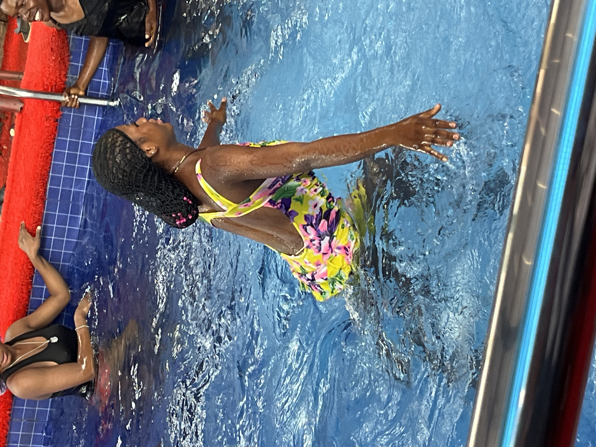 In a swimming pool located in Obosi, Nigeria, a Black woman with braided hair, wearing a black swim cap adorned with pink beads, is standing in the water. She is also wearing a yellow swimsuit with a floral pattern featuring pink and purple flowers. Her right hand is raised slightly, and her left hand is extended forward, touching the water. To her right, another Black woman, wearing a black tank top, is visible, holding onto a metal pole. The pool is filled with blue water, and the edge of the pool is lined with blue tiles and a red inflatable barrier.