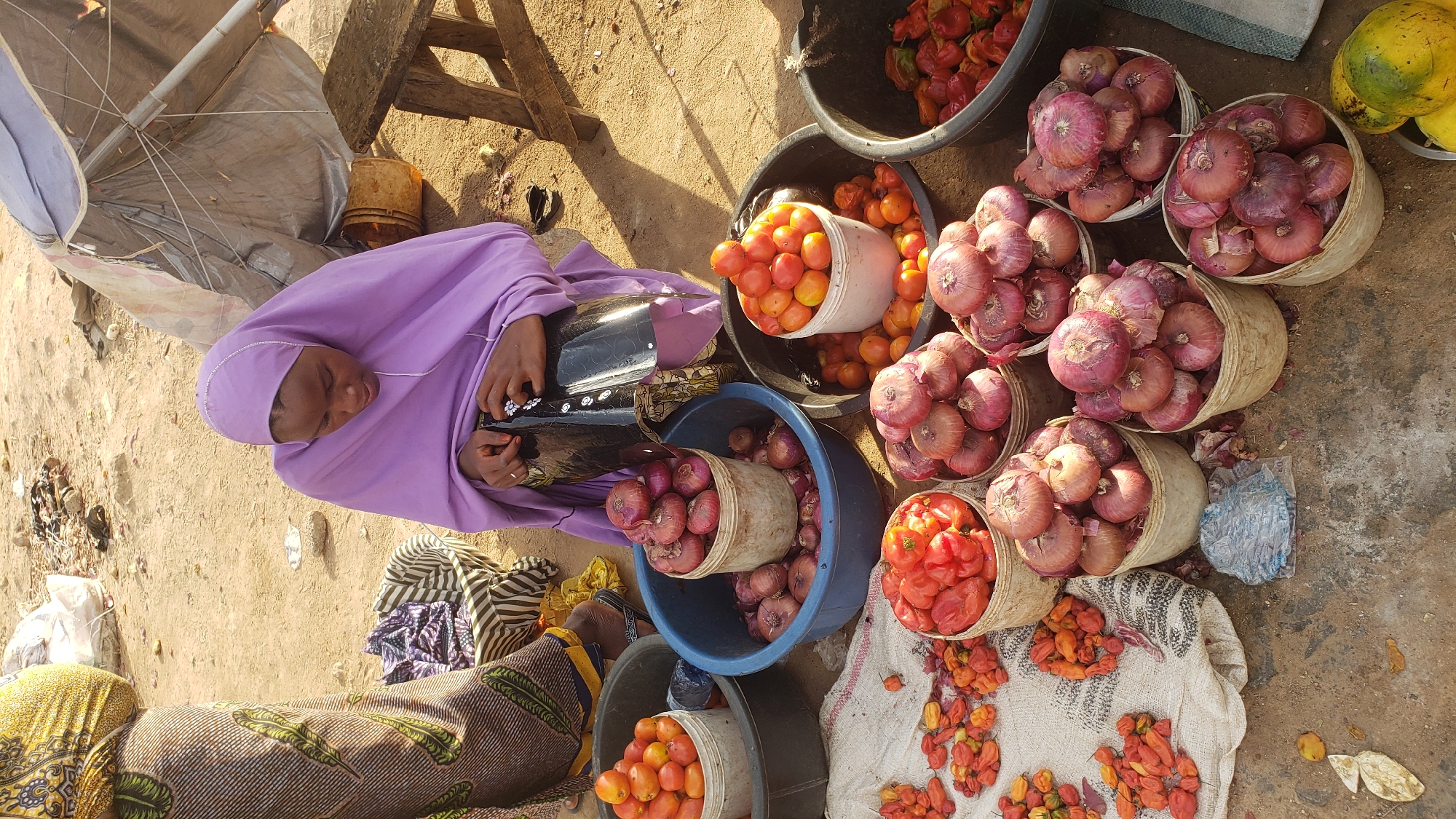 In Kawo, Nigeria, a woman wearing a purple hijab is sitting near an open-air market stall. She is positioned near buckets and baskets filled with produce including red onions, tomatoes, and peppers. An umbrella provides shade. Other individuals are partially visible in the foreground. The ground is sandy and shows signs of market activity.