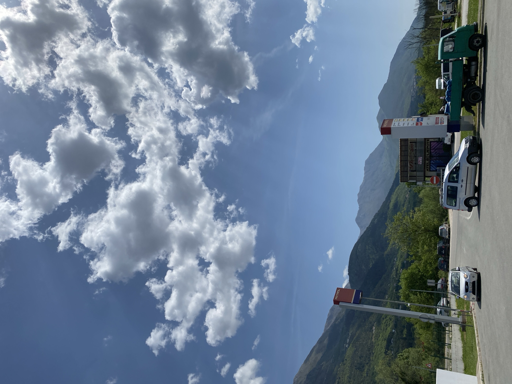 This image captures a bright, sunny daytime scene in Kavač, Montenegro, featuring a blend of natural mountainous landscape and a commercial roadside area. The sky is a prominent feature, displaying a clear blue canvas dotted with numerous fluffy white cumulus clouds of varying sizes, suggesting fair weather.
In the mid-ground, large, lush green mountains rise majestically, their slopes covered in trees and vegetation, indicating a vibrant natural environment.
The foreground and right side of the image show an asphalt road or parking lot, bustling with activity. A tall, multi-sided fuel station sign stands prominently, with one visible side displaying "PETROL" vertically in bold red letters against a white background. Below "PETROL," various price displays or service information panels are visible, though the text is not clearly legible. Behind this sign, other commercial structures can be seen, including a sign with "DREAM" written on it and another clearly stating "ATELJE LEPOTE" (Beauty Atelier/Salon).
Several vehicles are present: a white commercial van is positioned near a standard 'no entry' road sign, a small green open-bed truck is parked further right, and a white hatchback car is partially visible in the lower right. Additional cars are parked in the background, suggesting a busy stop. The bright sunlight and relatively short shadows indicate it is likely late morning or early afternoon. The scene depicts a functional roadside stop nestled within a scenic mountainous region.