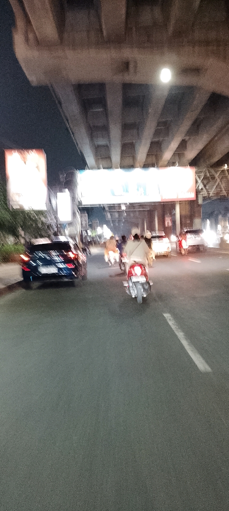 The image depicts a street scene at night in Bekasi, Indonesia, likely under an elevated highway structure. The foreground is dominated by a dark asphalt road with faint white lane markings. Several vehicles are visible, including a dark blue car on the left, a red scooter with a rider in the center, and other motorcycles and cars further down the road, all of which are blurred due to motion. There are also several figures on motorcycles, their forms indistinct due to motion blur. Overhead, the concrete supports and beams of an elevated structure are prominent, casting shadows and creating a darkened environment. Brightly lit billboards or signs are visible in the background, suggesting an urban commercial area. The overall impression is one of nighttime traffic congestion in a city.