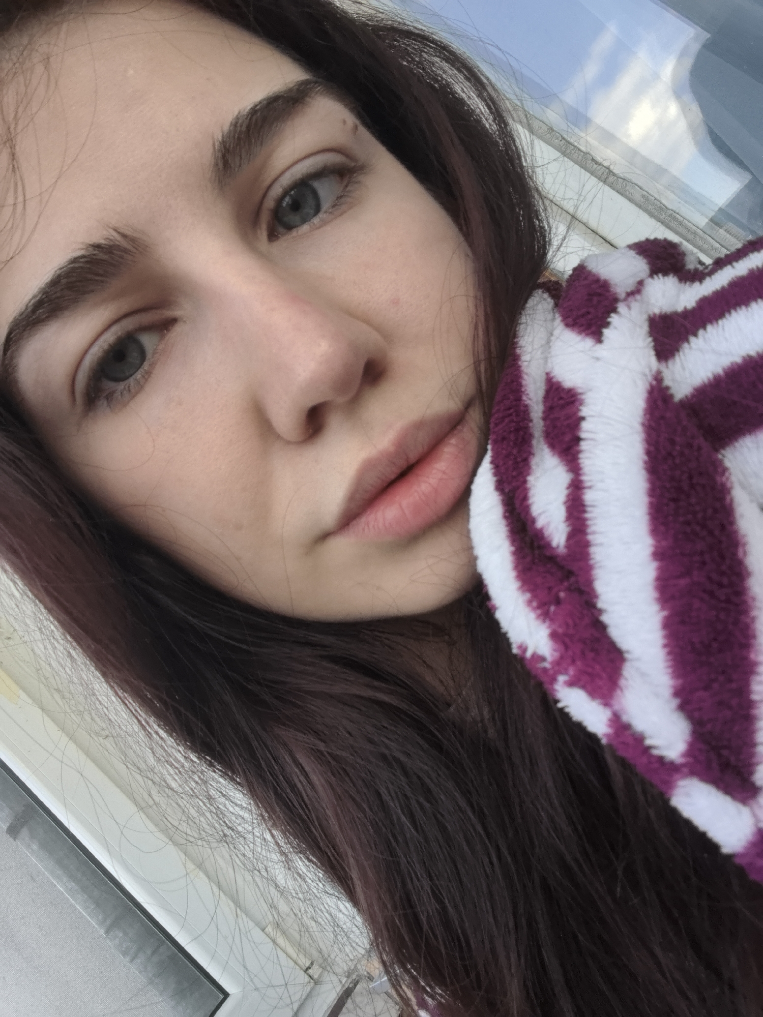 A close-up selfie of a young woman with long, auburn-brown hair and light, possibly grey eyes. She has fair skin and full lips. The woman is holding a fluffy towel with purple and white stripes up to her face on the right side of the frame. A window frame is visible in the background. It is not possible to determine from the image if the location is Stara Zagora, Bulgaria.
