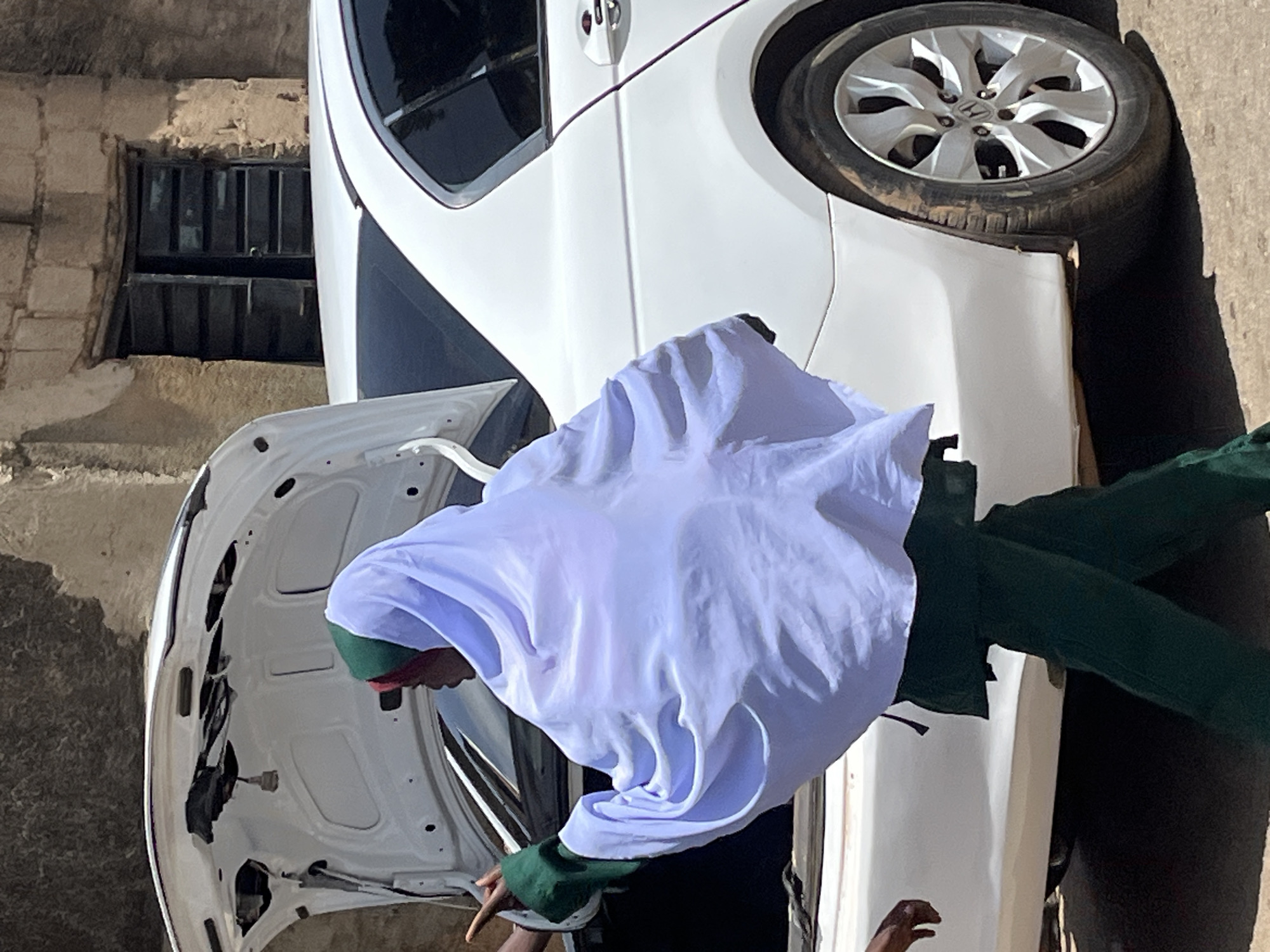 In Jos, Nigeria, a person wearing a white hijab and green clothing is seen from behind, standing next to a white car with its trunk open. The person's right arm is extended, and their hand is pointing towards something out of frame. Another person's hand is visible reaching towards the person in the hijab. The background consists of a stone building with a dark, wooden door and window frames. The scene appears to be outdoors on a sunny day.