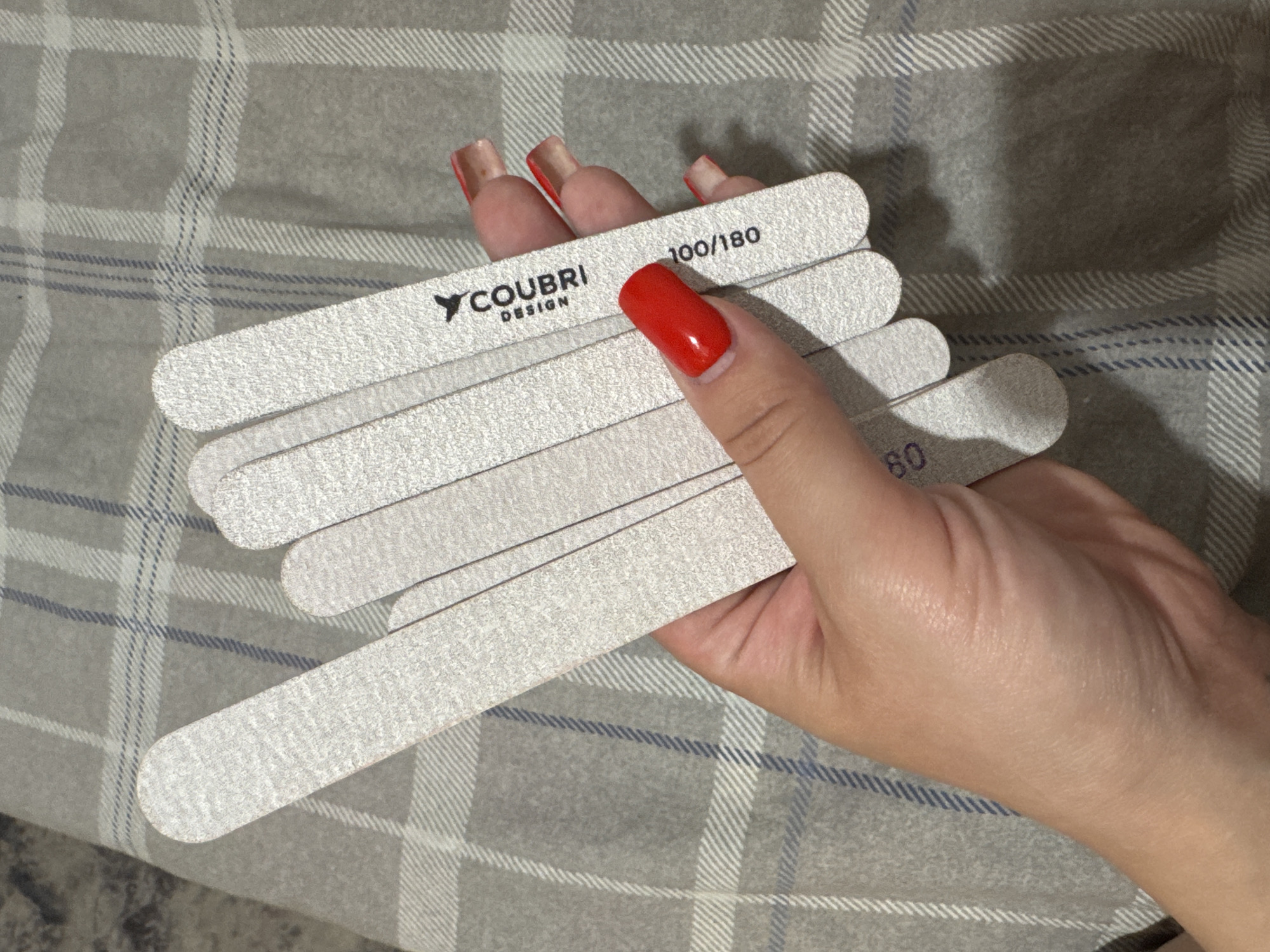 A hand with red painted fingernails holds a fan of six white nail files against a grey and white checkered background. The nail files are arranged in a fanned-out pattern, with the widest part towards the bottom. The hand is holding the files in the center, with the thumb on top and the fingers underneath. The checkered background appears to be a blanket or fabric. There is a logo that reads "COUBRI DESIGN" on one of the nail files. Another nail file has the numbers "100/180" printed on it, indicating its grit. A nail file on the far left has the number "80" printed on it. There are no other people or objects visible in the frame. The lighting is moderate, suggesting it is indoors.