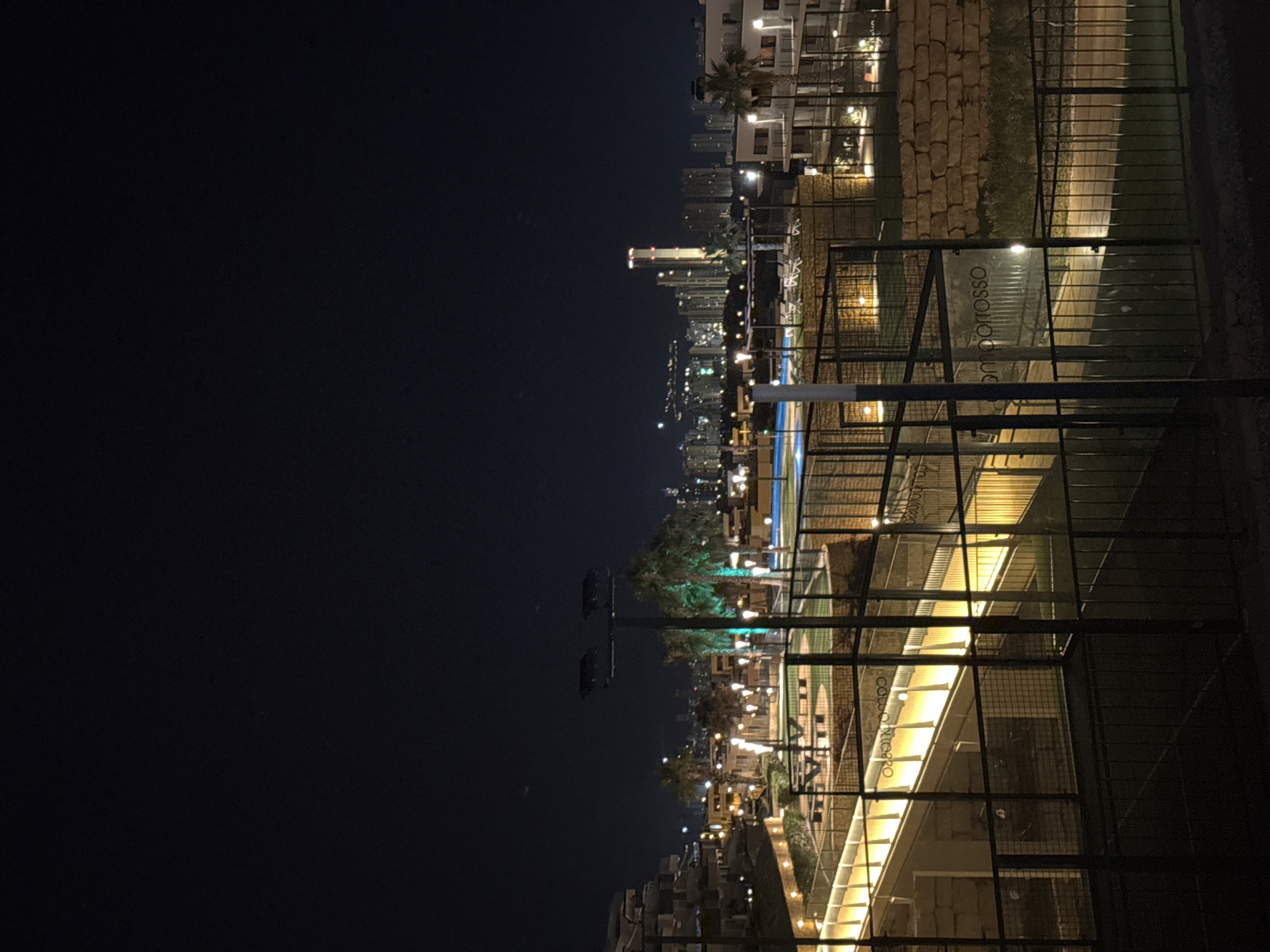 The image captures a nighttime view of a resort or residential complex, likely in Finestrat, Spain, as indicated by the presence of palm trees and a coastal city skyline in the distance. The foreground is dominated by a black metal fence, obscuring a sports court, possibly padel tennis, given the context of Spain. Beyond the fence, a brightly lit walkway with yellow lights runs diagonally upwards. Further back, there are illuminated palm trees with green lights, a swimming pool, and several buildings with visible lights. The background features a sprawling city skyline with numerous buildings, including a prominent tall tower with illuminated red accents at its peak. The overall atmosphere is one of a modern, illuminated leisure and residential area at night.