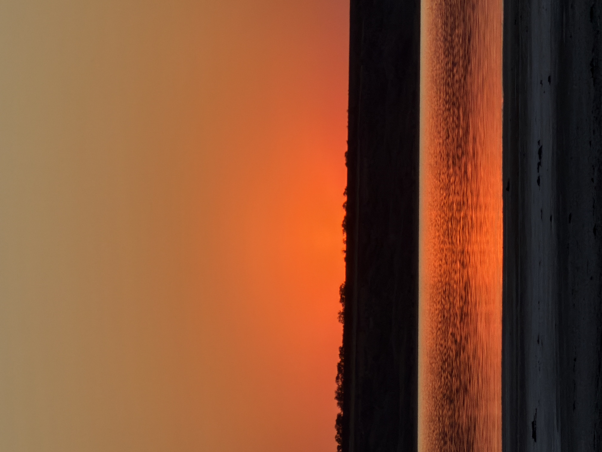 The image depicts a tranquil sunset scene over a body of water, likely a lake or a wide river, in the village of Korsuntsi, Ukraine. The sky is a vibrant gradient of orange and amber, transitioning to a lighter tan hue at the top of the frame. A dark, silhouetted horizon line separates the sky from the landscape below, with a sparse line of trees visible along its edge.
The water's surface reflects the intense colors of the sunset, creating a shimmering, textured expanse of orange light. In the foreground, a dark, sandy or muddy shoreline is visible, with patches of lighter, possibly salt-encrusted earth. The overall atmosphere is serene and peaceful, with no human presence or activity depicted. The scene is captured from a slightly low vantage point, emphasizing the vastness of the sky and the reflective quality of the water. The time of day is clearly sunset, characterized by the intense coloration of the sky and its reflection.