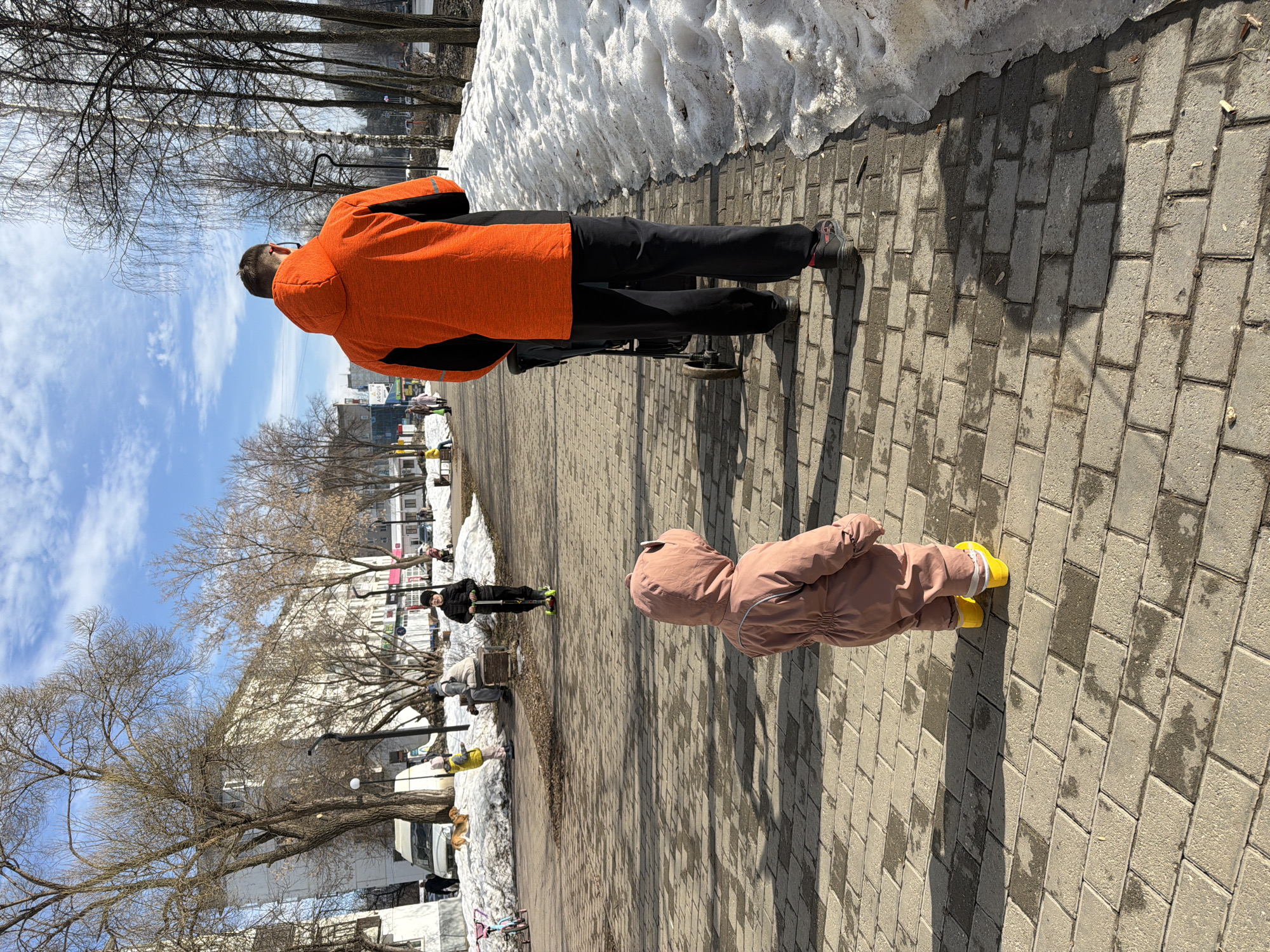 This image depicts a park or pedestrian promenade in Chaikovsky, Russia, during what appears to be a sunny day in late winter or early spring. The scene is illuminated by strong sunlight under a clear blue sky with scattered white clouds.
In the foreground, a young child, viewed from the rear, stands on a damp, gray paver walkway. The child is dressed in a pink hooded full-body suit featuring small ear-like protrusions on the hood, paired with bright yellow boots. To the child's right, an adult, also viewed from the rear, walks along the same walkway, pushing a black baby stroller. This individual wears an orange and black hooded jacket and black pants.
The right side of the walkway is lined with large, dirty piles of melting snow. Further down the path, a child in a black jacket and hat rides a scooter equipped with green wheels. In the background, a child wearing a yellow jacket is observed near a snow pile where a brown dog is lying down. Another adult is seated on a bench beside a snowdrift. Numerous bare deciduous trees, including some with light-colored bark, are scattered throughout the park area and line the background. Beyond the trees, multi-story, light-colored buildings are visible, lining what appears to be a street, with a white van parked near one of the structures. Distinct shadows of the subjects and trees are cast upon the wet paver surface.