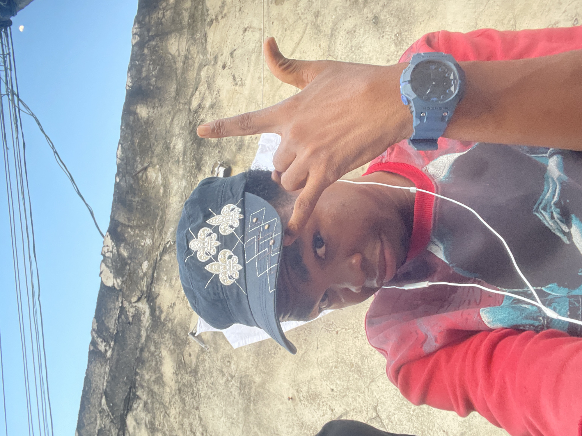 The media file is a close-up, low-angle, portrait-style photograph taken outdoors, likely in Ado, Nigeria. The main subject is a person, whose face is partially visible, looking towards the camera. They are wearing a dark baseball cap adorned with white floral embroidery and a diamond-shaped pattern. They also have on a red long-sleeved shirt. Earbuds with a white wire are visible, suggesting the person is listening to audio.
A hand is prominently featured in the foreground, extended towards the camera, with two fingers forming a "V" shape, often interpreted as a peace sign or gesture of victory. The person is also wearing a blue digital watch on their wrist. The background consists of a rough, textured, light-colored wall and a clear blue sky. The lighting suggests it is daytime, likely mid-morning or afternoon. The image has a casual, candid feel. There is no visible text in the media file.