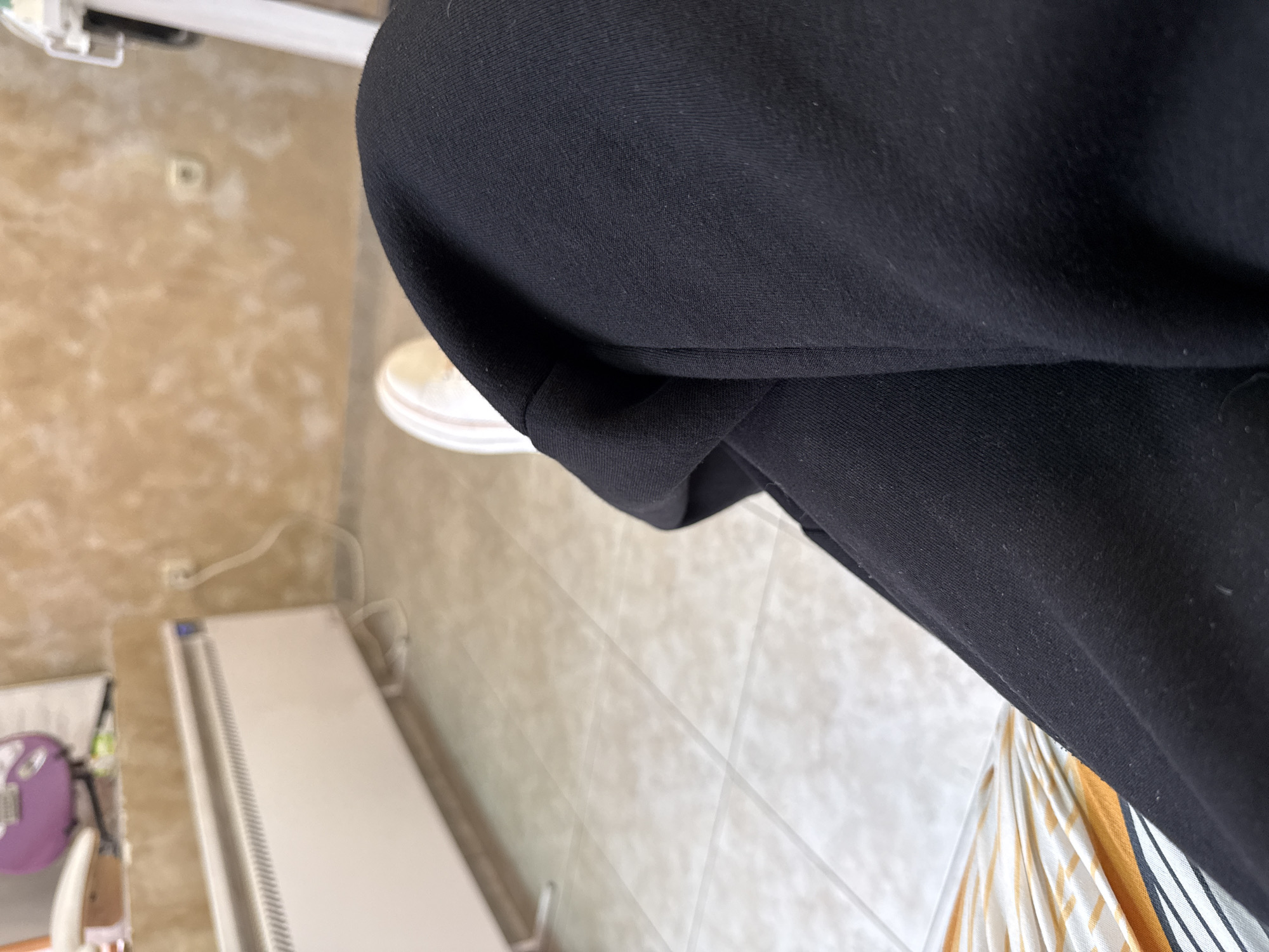 The image presents an indoor scene. The foreground is dominated by a black garment, possibly a piece of clothing, that is out of focus and close to the camera. A white shoe peeks from behind the garment. In the mid-ground, the floor is tiled in a light neutral tone. To the left, there's a blurry view of a white radiator and what appears to be a table or furniture piece with a purple object on it. The background shows a textured wall in a beige or light brown hue, with an electrical outlet and a visible power cord. The photo doesn't provide enough context to determine a specific location within Sofia, Bulgaria. There are no visible people, actions, or interactions depicted in the image.