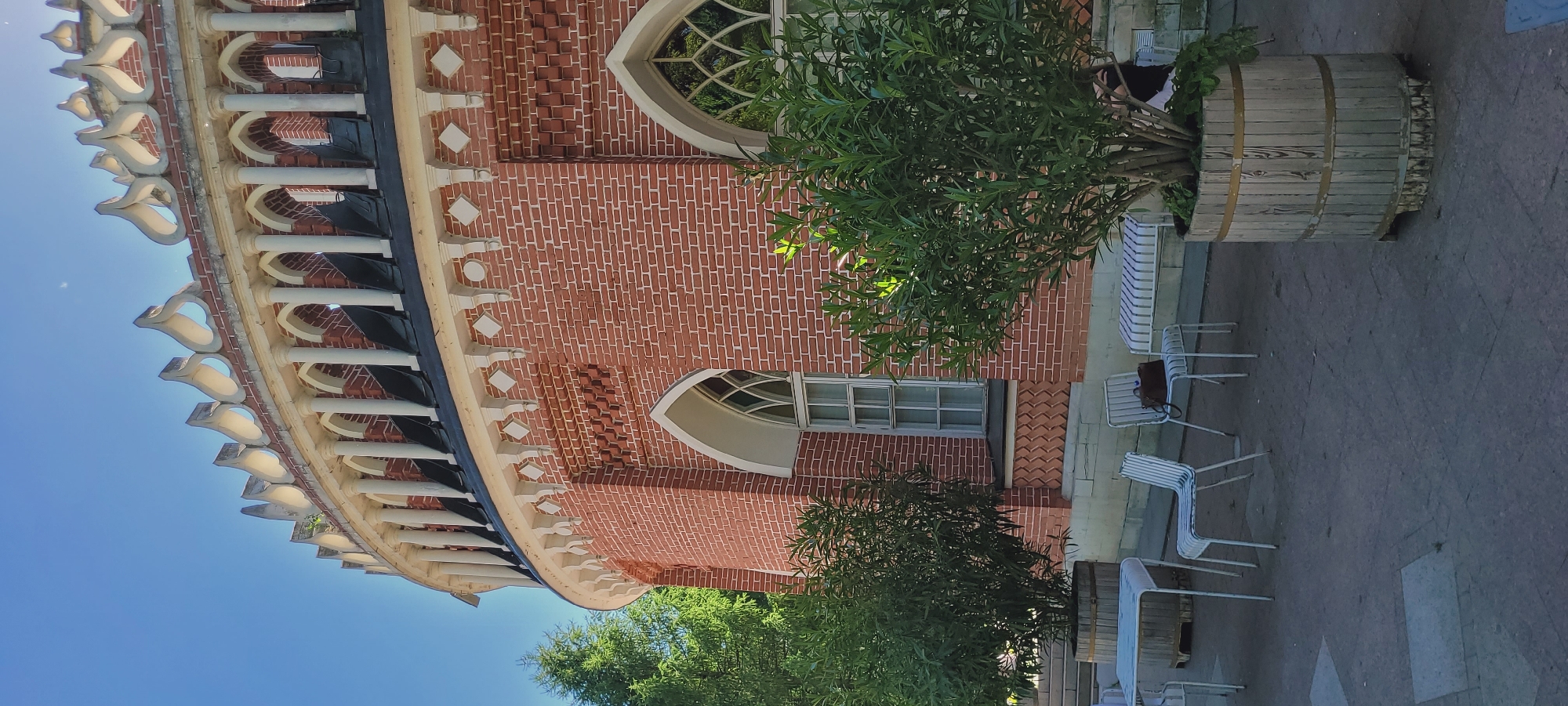 The image displays a detailed view of a distinct red brick building, characteristic of the **Tsaritsyno Museum-Reserve in Moscow, Russia**. The building features a curved facade with numerous architectural details. On the lower level, white-framed Gothic-arched windows are set into the red brickwork, which includes decorative patterns such as recessed rectangles and a zig-zag motif below the window sills. A white cornice adorned with corbels, circular, and diamond-shaped elements runs along the top of this section. Above, a darker band precedes an open arcade composed of slender white columns. The structure culminates in an ornate white parapet featuring a repetitive, flame-like or heart-shaped decorative motif, silhouetted against a bright, clear blue sky.
In the foreground, an outdoor seating area is visible on a grey tiled patio. It includes a white metal square table and several matching white slatted chairs; a dark brown handbag rests on one of the chairs. A white and light blue/grey striped bench is also present. Several large green shrubs, possibly Oleander, are planted in light-colored wooden barrel planters, partially obscuring the building's base. Taller green trees are visible to the far left. One person with dark hair is partially visible, seated behind a large potted plant on the right side of the frame. The scene is brightly lit by natural sunlight, casting subtle shadows and indicating a clear day.