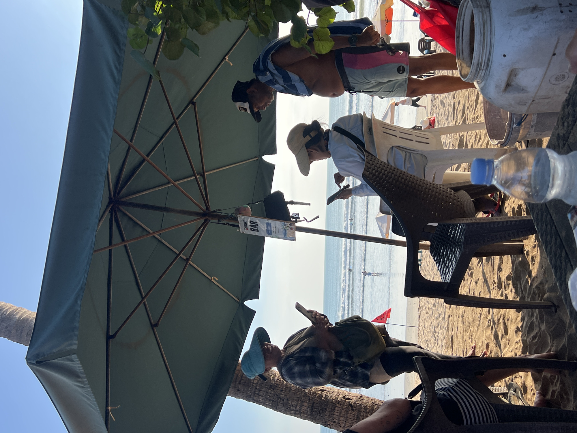 The image captures a scene on a beach in Kuta, Indonesia, under a large green umbrella providing shade. Three individuals are visible in the foreground. On the left, a woman wearing a blue wide-brimmed hat and a plaid shirt is looking at her phone. In the center, a person in a white shirt and a light-colored hat is also engrossed in their phone, seated in a white plastic chair. To the right, a man with a towel draped over his shoulders and wearing swim trunks stands, looking downwards. The background reveals the ocean with gentle waves and a few people in the water, suggesting a typical beach day. The sand is golden, and a palm tree trunk is visible on the left. Several plastic chairs are arranged under the umbrella, along with a large white container and a bottle of water on a table in the immediate foreground. A sign for "CHERRY BAR" is attached to a pole near the umbrella. The overall atmosphere is relaxed and casual, indicative of a beachside establishment.