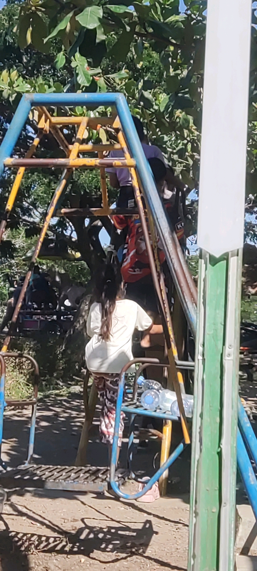 In Gebang Kulon, Indonesia, a group of children are gathered around a weathered metal playground structure. The structure, painted in blues and yellows, appears to be a swing set and climbing frame, with rusted supports and worn platforms. One child, a girl with long dark hair, is seen from behind, wearing a light-colored t-shirt and patterned pants, as she climbs the structure. Other children are partially visible, some higher up on the frame and others in the background. The ground beneath the playground is a dirt and gravel mix, with shadows cast by trees and the playground equipment. Several plastic water bottles are scattered on a lower platform of the structure. The scene is set outdoors, with dense green foliage and tree branches overhead, indicating a natural, park-like environment.
