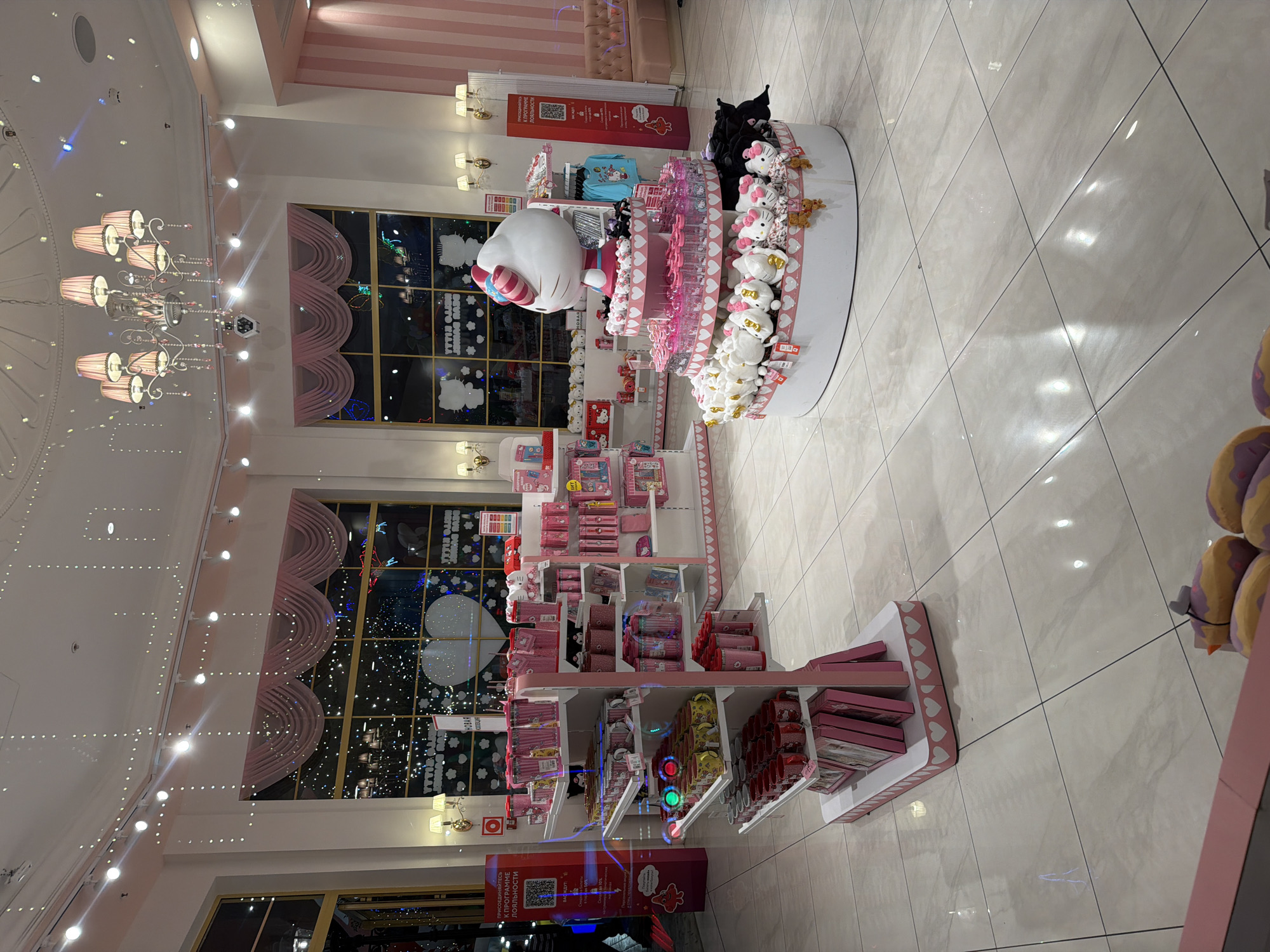 The image shows the interior of a brightly lit store, decorated in pink and white, with a large display of Hello Kitty merchandise. In the center of the store, a multi-tiered circular display stand is prominently featured. The stand showcases a large Hello Kitty figurine at the top, surrounded by an assortment of plush toys, water bottles, and other items. The lower tiers of the stand are filled with white and black plush toys, as well as pink water bottles.
To the left of the central display, a white retail shelving unit is filled with pink-themed merchandise, including mugs and cups. The shelves are organized with various sizes and types of items, all in shades of pink. Further to the left, a tall, narrow display stand with red signage is visible, and a person in dark clothing is partially visible walking past.
In the background, large windows are visible, displaying a nighttime scene with illuminated decorations, including a large heart shape. Pink curtains with string lights are draped across the top of the windows. The ceiling features a large, ornate pink chandelier and numerous small string lights. The floor is tiled with large, pale beige tiles. On the right side of the store, another shelving unit with pink merchandise is partially visible, along with a pink-striped wall and a portion of a pink upholstered seating area. The overall atmosphere is festive and designed to appeal to fans of the Hello Kitty brand. The location is Unknown, Unknown.