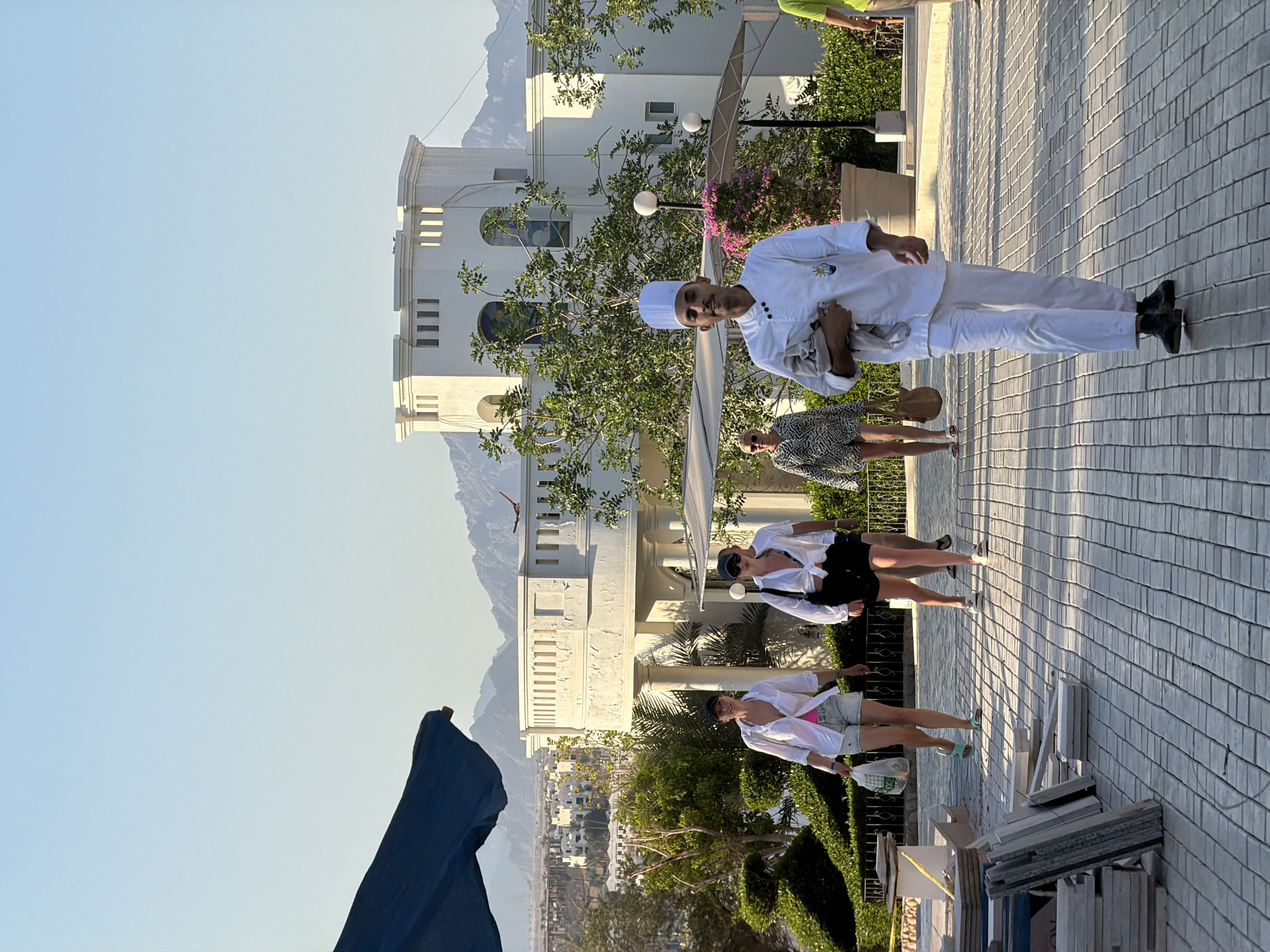 The image depicts a sunlit outdoor scene, likely in Sharm El Sheikh, Egypt. A man in a white chef's uniform and hat is walking towards the viewer, holding a folded grey garment. He has a light-colored face and dark hair. Behind him, three women are walking away from the viewer. The woman closest to the chef is wearing a black top and black shorts. To her left, a woman in a white shirt and denim shorts is carrying a white plastic bag. Further left, a woman in a patterned dress is walking. In the background, a large white building with a turret-like structure and multiple windows is visible, with mountainous terrain beyond it. Lush green foliage and flowering plants are scattered throughout the scene. The ground is paved with light-colored rectangular tiles. A dark blue awning or umbrella is partially visible in the upper left corner.