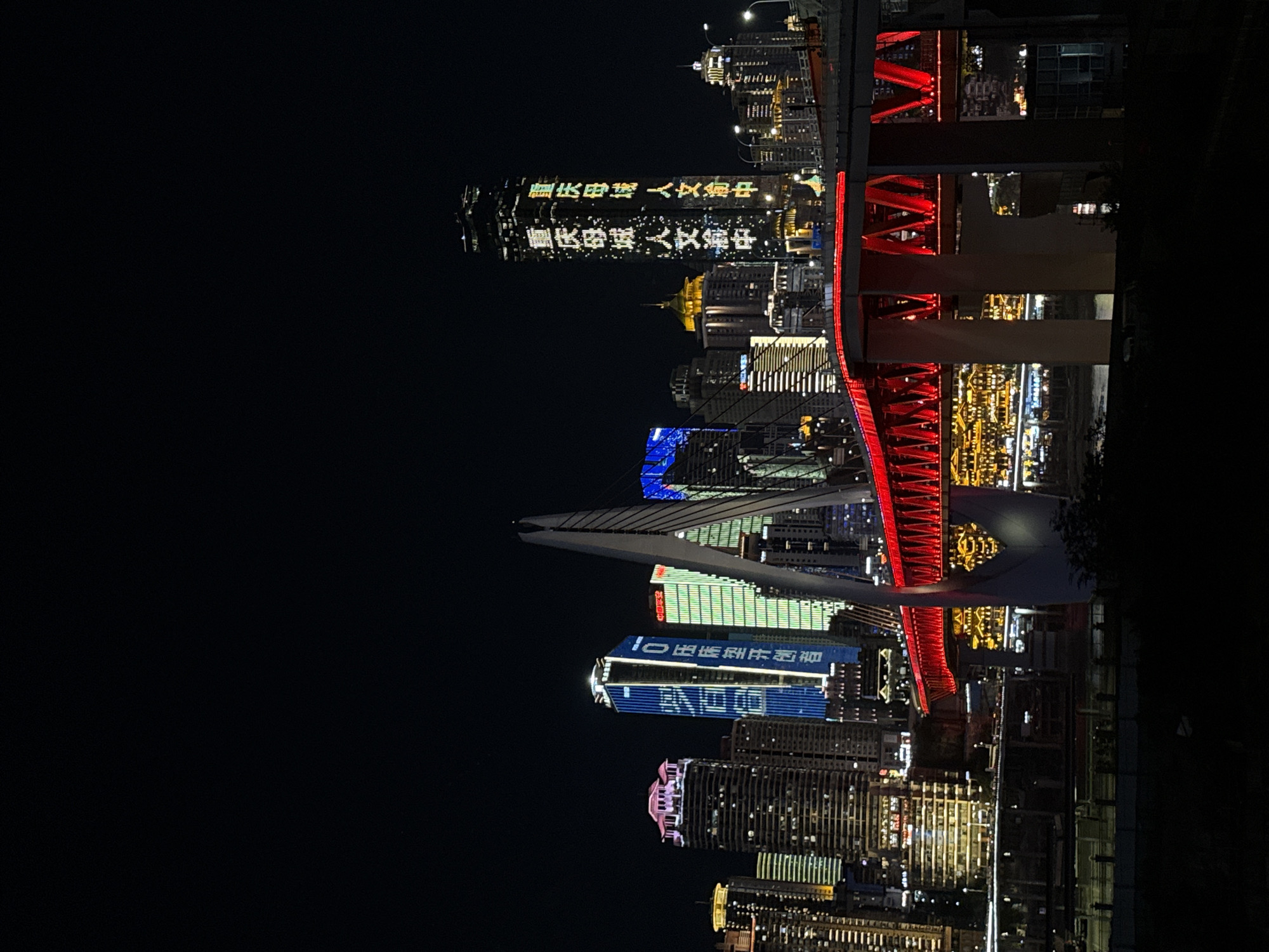 A nighttime, wide-angle, low-angle shot captures a vibrant cityscape dominated by illuminated skyscrapers and a prominent, brightly lit red bridge. The scene, located in the Jiangbeicheng Street area of China, features a dark sky that contrasts sharply with the array of lights emanating from the buildings and infrastructure.
In the foreground, a multi-tiered red bridge with visible supporting structures stretches across the frame, its vibrant illumination drawing the viewer's eye. Behind the bridge, a dense collection of modern skyscrapers rises, their facades adorned with various colors and patterns of light. One particularly tall, slender skyscraper displays text in glowing green and yellow characters. Other buildings showcase blue and white illuminated text, as well as solid blocks of color like pink and green, highlighting their architectural features. Smaller buildings with warm, golden lights are visible in the mid-ground, suggesting a busy urban environment.
The background is a deep black, indicating a clear night sky, with no celestial bodies or atmospheric phenomena visible. The overall impression is one of a dynamic and technologically advanced urban center, alive with light and activity after dark. There are no people or vehicles clearly discernible in the image, emphasizing the architectural and environmental aspects of the scene. The image is devoid of any foliage or natural elements, reinforcing the urban setting.