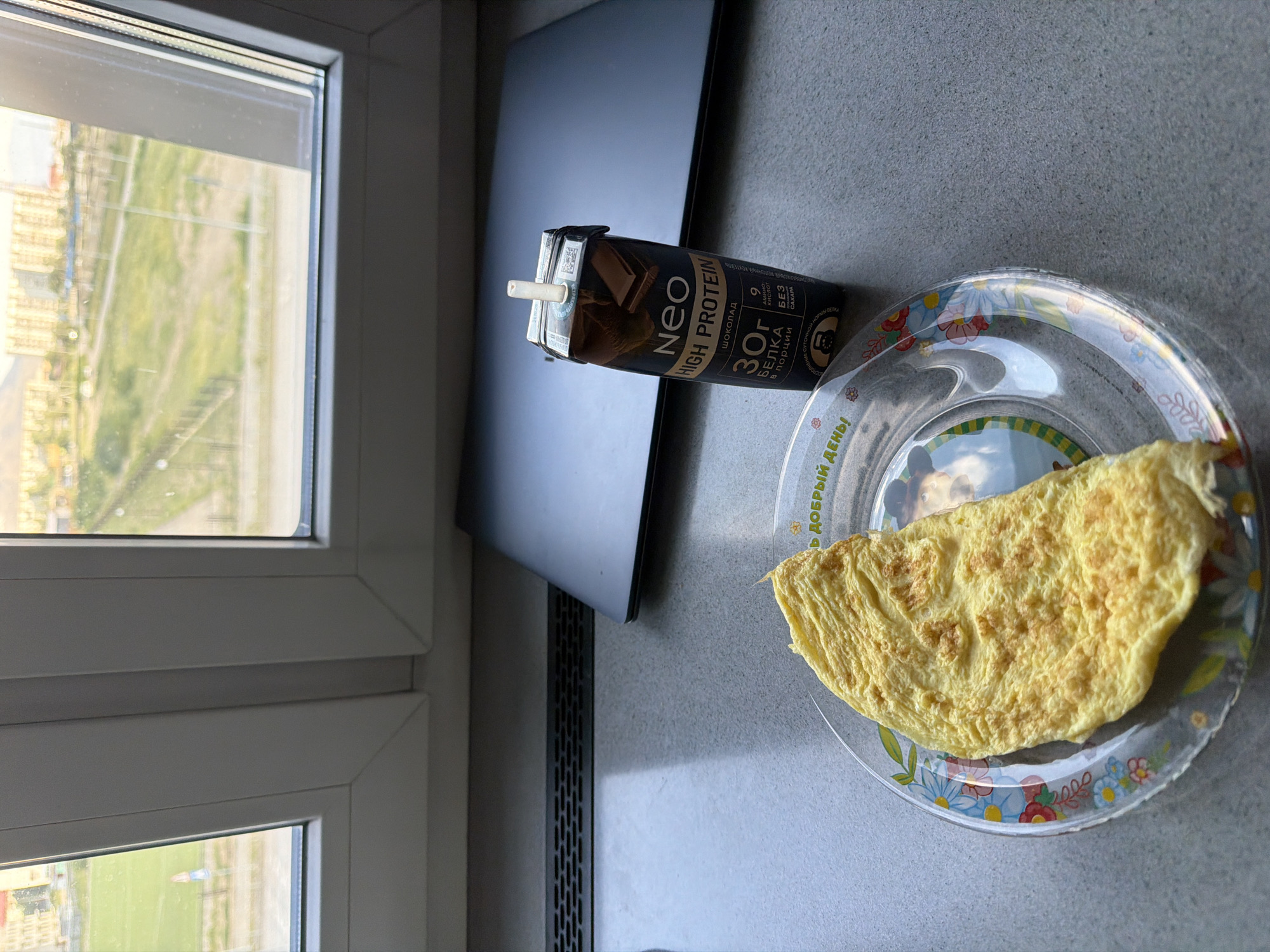 An overhead shot captures a breakfast scene on a gray countertop, positioned on a windowsill overlooking a city. A fluffy, golden-yellow omelet rests on a clear plastic plate adorned with a floral pattern. To the right of the plate sits a dark-colored carton of "Neo High Protein" chocolate drink, its straw extended. Behind these items, a closed laptop is visible, its dark surface reflecting light. The background reveals a view of buildings and green spaces through a window, suggesting an urban environment. The overall lighting is soft, indicating it might be daytime. The specific location is identified as Kyzyl, Russia.