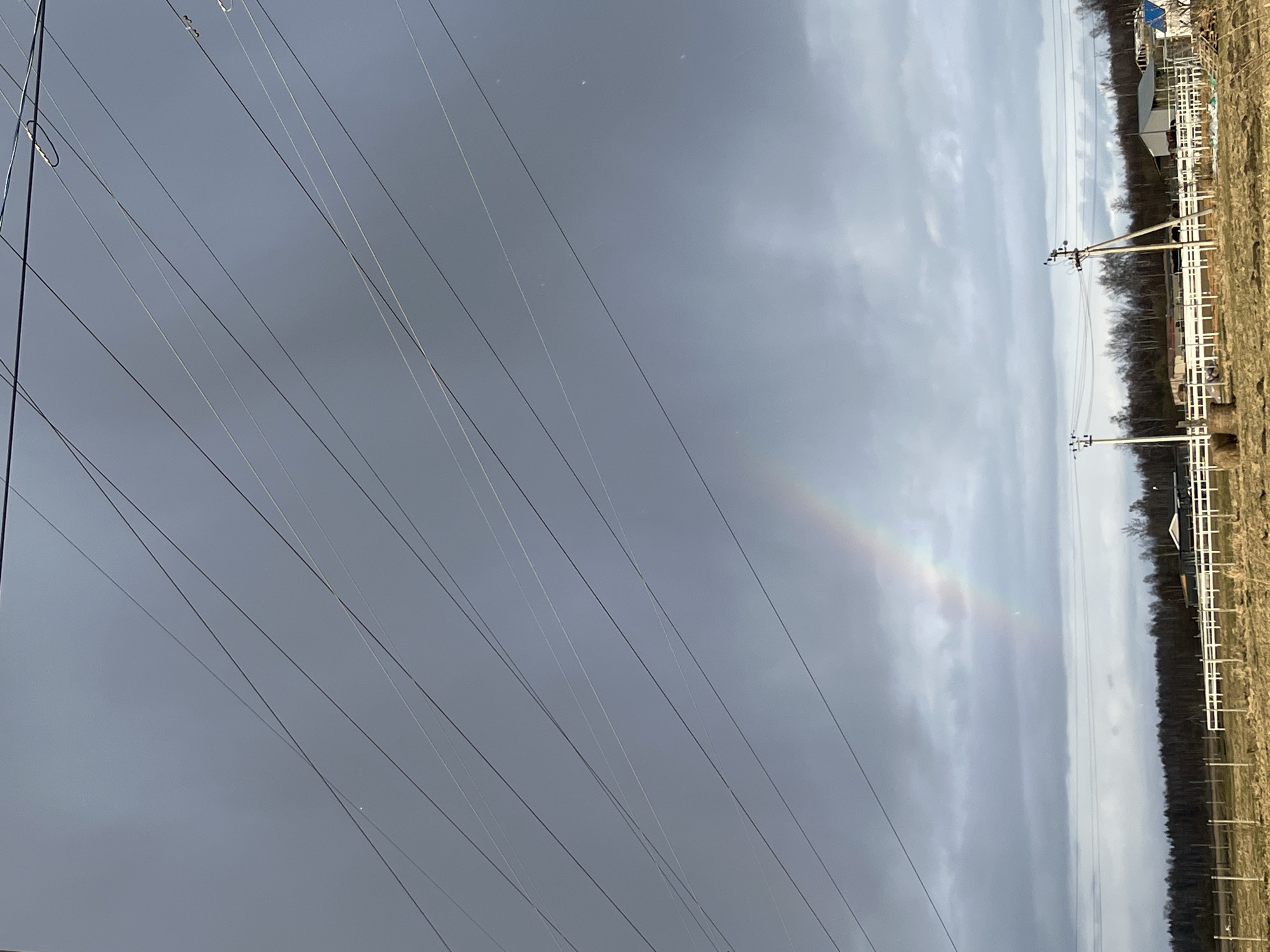 The image depicts an overcast sky in Razdol'e, Russia, bisected by numerous power lines. A faint rainbow is visible in the center of the frame, arching from the horizon upwards. On the right side of the image, there is a grassy field and a white fence line leading to some buildings and trees. Electrical poles with transformers are placed alongside the fence.