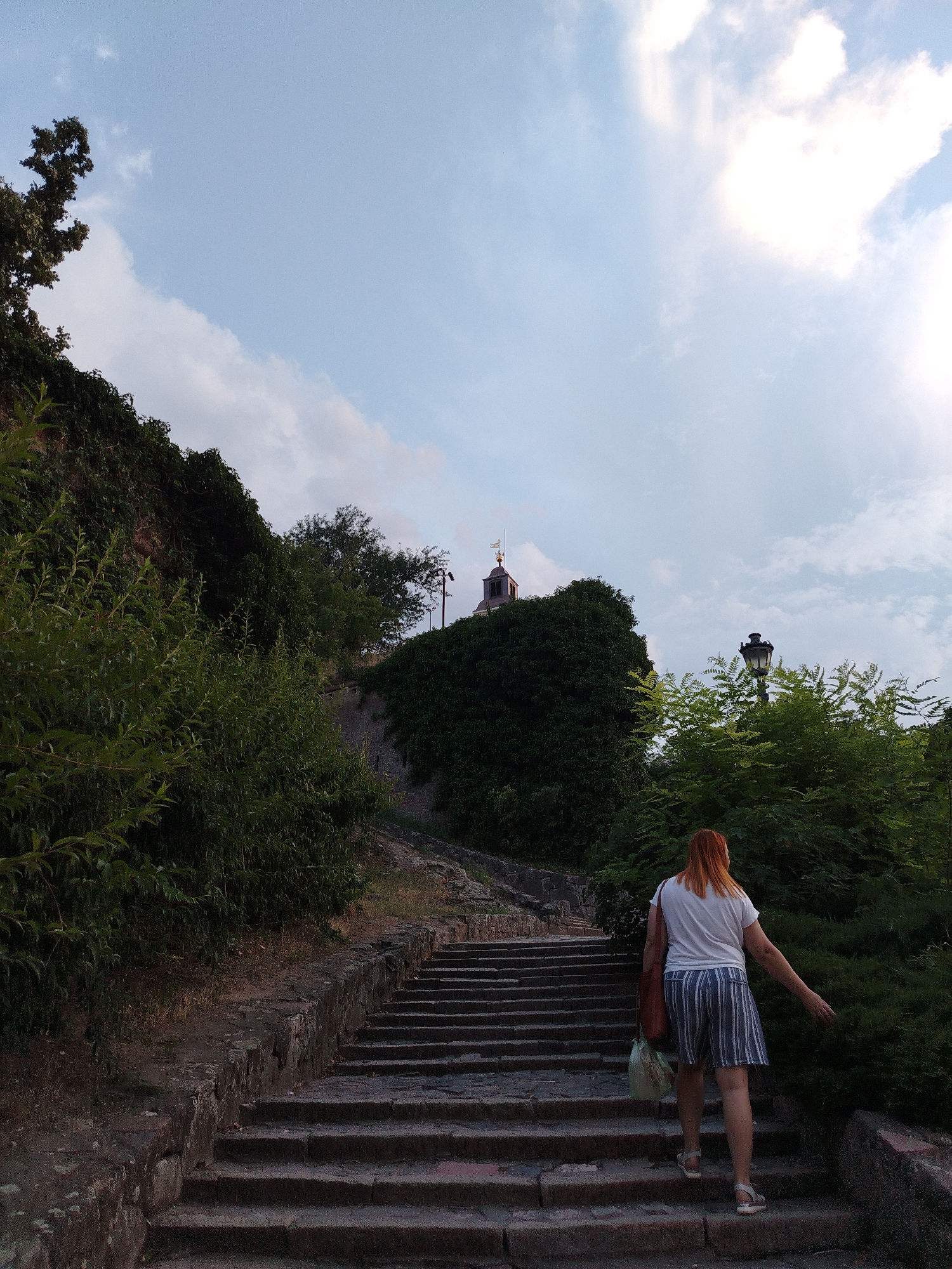 The image captures a view of the Petrovaradin Fortress in Petrovaradin, Serbia. A woman with reddish-blonde hair is ascending a stone staircase, her back to the camera. She wears a white t-shirt and striped shorts, and carries a brown bag over her shoulder and a light-colored plastic bag in her left hand. Lush green foliage lines the sides of the staircase and the surrounding terrain. In the background, atop the fortress's ramparts, a clock tower with a dark roof and a weather vane is visible, partially obscured by trees and greenery. A black lamppost stands to the right of the tower. The sky above is a pale blue with scattered white clouds.