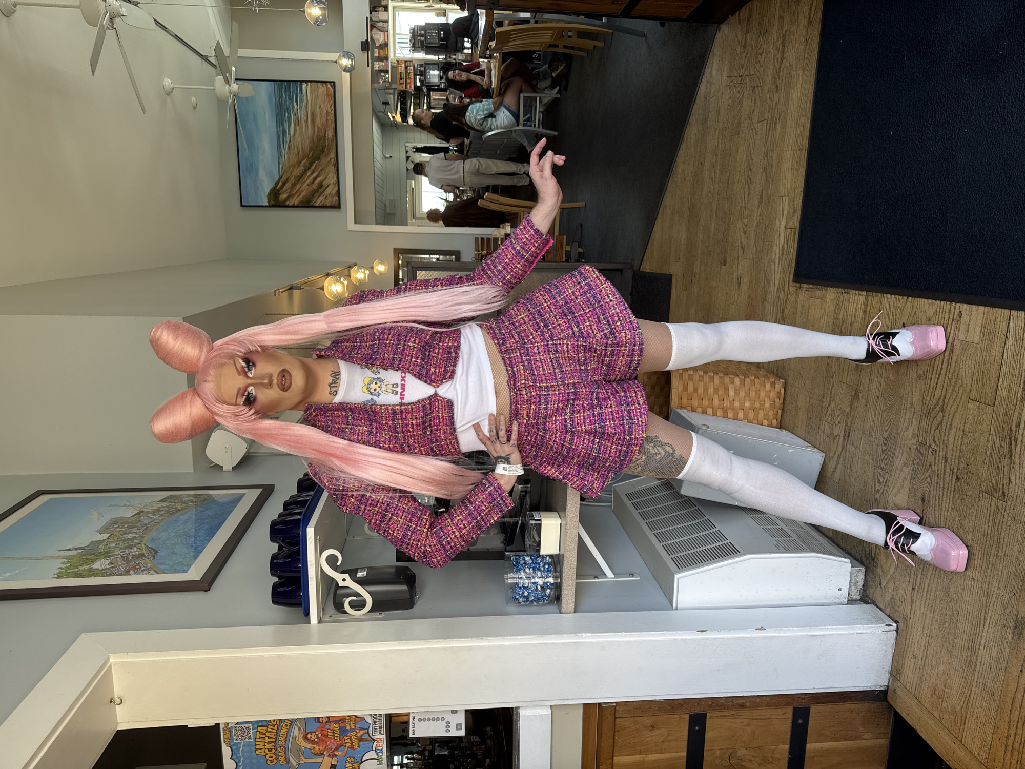 A drag performer with long, pink, cartoon-like hair styled in two buns stands in a split in what appears to be a restaurant or bar. The performer wears a pink tweed suit jacket and matching skirt, a white t-shirt with a cartoon graphic, white thigh-high stockings, and pink and black spectator shoes. The performer has heavy makeup and tattoos on their arms and legs.
The scene is indoors, with wood-paneled flooring and light-colored walls. A framed painting of a coastal scene hangs on the wall. In the background, other patrons are visible seated at tables, suggesting a public space. A sign with the words "ANITA COCKTAIL'S DRAG BRUNCH" and "BAR SEATING" is visible at the bottom of the frame, providing context for the event. The overall lighting suggests daytime.