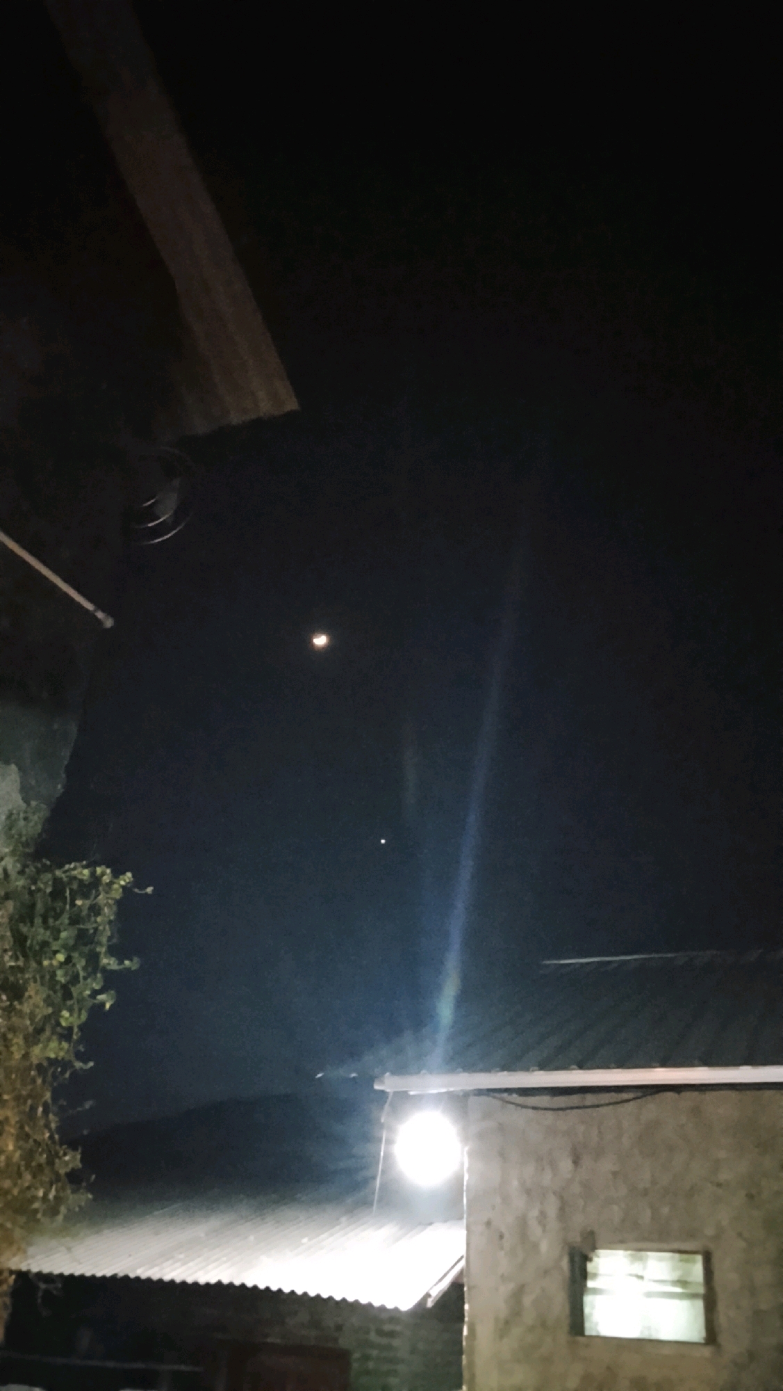 The image depicts a nighttime scene in Ujungrusi, Indonesia, featuring a dark sky with a crescent moon visible. Below the moon, a smaller celestial body, possibly a planet, is also discernible. In the foreground, a corrugated metal roof covers what appears to be a building with a concrete wall. A bright outdoor light illuminates the roof and part of the wall, creating a strong glare. To the left of the building, a portion of a structure with an overhead wooden beam is visible. Lush green foliage is also present on the left side of the frame, extending upwards. The overall impression is one of a rural or semi-urban residential area at night.