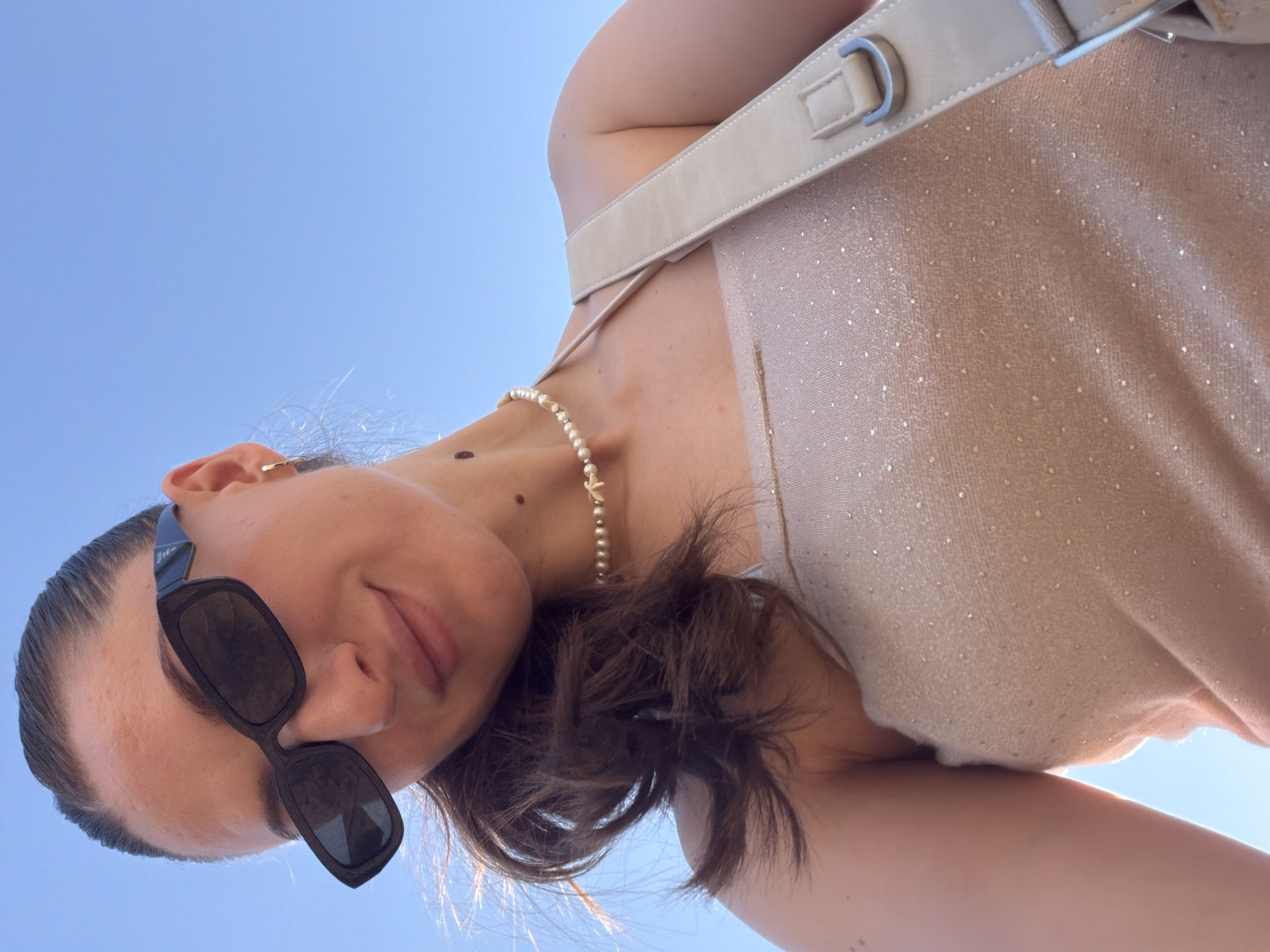A woman with dark hair pulled back in a ponytail, wearing black rectangular sunglasses, a pearl necklace with a starfish pendant, and a sparkly beige strapless top, is pictured from a low angle against a bright blue sky. A light beige purse strap is visible across her shoulder. The background appears to be a clear sky, suggesting an outdoor setting. The image does not contain any information to indicate the location is Sharm El Sheikh, Egypt.