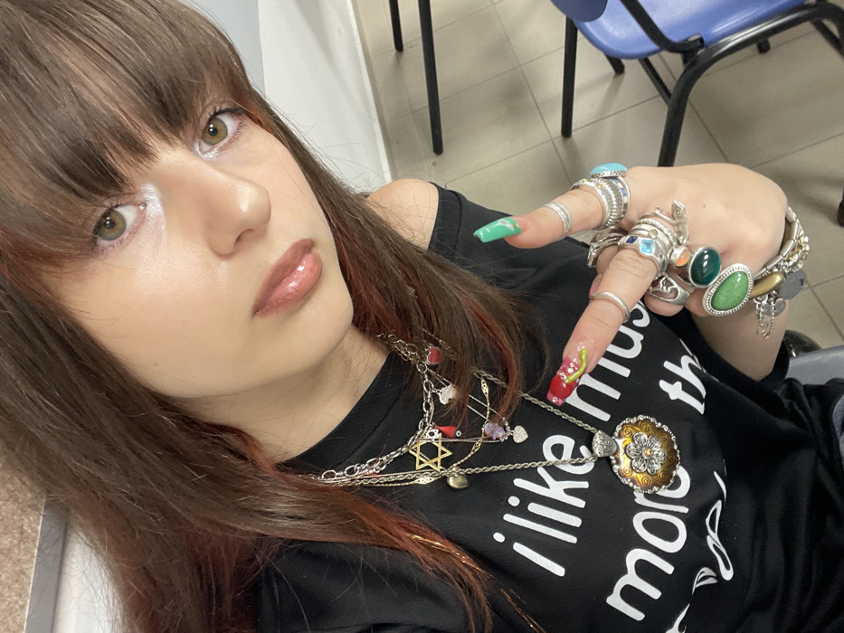 A young female subject is depicted in a close-up, selfie-style photograph taken indoors in Raanana, Israel. She has long, dark brown hair with reddish highlights and bangs, and light-colored eyes. Her skin appears fair, and she is wearing light makeup including eyeshadow and lip gloss. She wears a black, off-the-shoulder shirt with partially visible white text reading "i like more th". Multiple layered silver-tone necklaces are visible, one featuring a Star of David pendant and another a large, circular, ornate pendant with a central floral design in gold and brown tones. Various smaller charms, including hearts, are also present on the necklaces. Her left hand is raised, displaying numerous rings on most fingers, featuring various designs including plain bands and rings set with green, blue, and turquoise stones. Her fingernails are long and elaborately decorated: the index finger has a red base with green stem/leaf accents, and the middle finger has a green base with white swirl patterns. The background consists of a plain light-colored wall and the partial view of a dark-framed chair with a blue seat.
