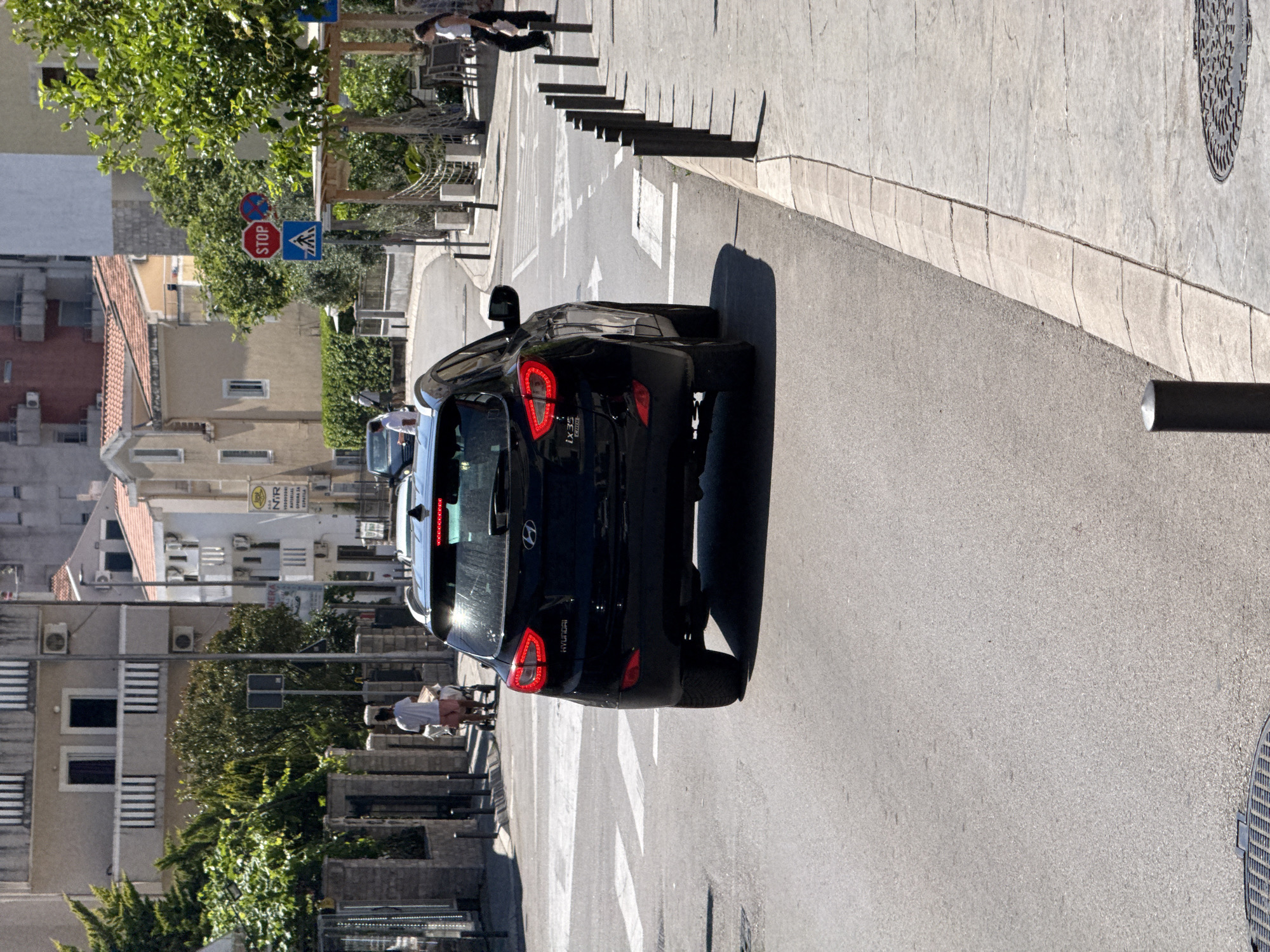The image depicts a black Hyundai ix35 SUV driving on a street in Budva, Montenegro. The vehicle is in motion, approaching a pedestrian crossing. In the background, there are buildings with typical Mediterranean architecture, including tiled roofs and balconies. Several trees and greenery are visible, suggesting a well-maintained urban environment. A stop sign and a pedestrian crossing sign are prominently displayed at an intersection ahead. People are seen on the sidewalks, including a person pushing a stroller and another walking. The overall scene conveys a sense of everyday urban life in a coastal town.
