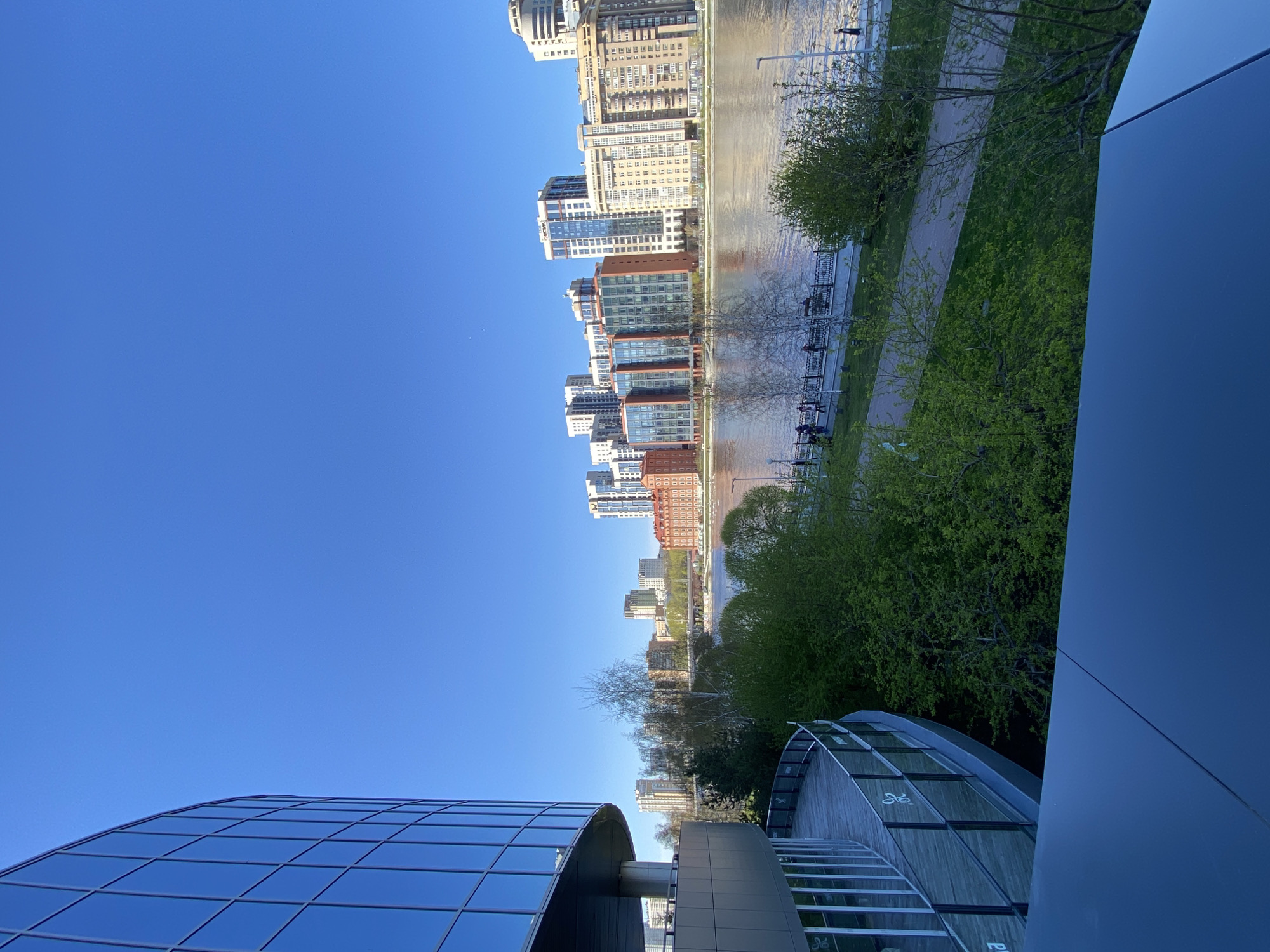 The image shows a cityscape in Yekaterinburg, Russia, viewed from a high vantage point, likely atop a building. A river bisects the scene, with modern high-rise buildings on the opposite bank. The riverbank is lined with a pedestrian walkway, trees, and some benches. The foreground features parts of a modern architectural structure, including a glass-domed roof and a smooth, solid wall. The sky is clear and blue.