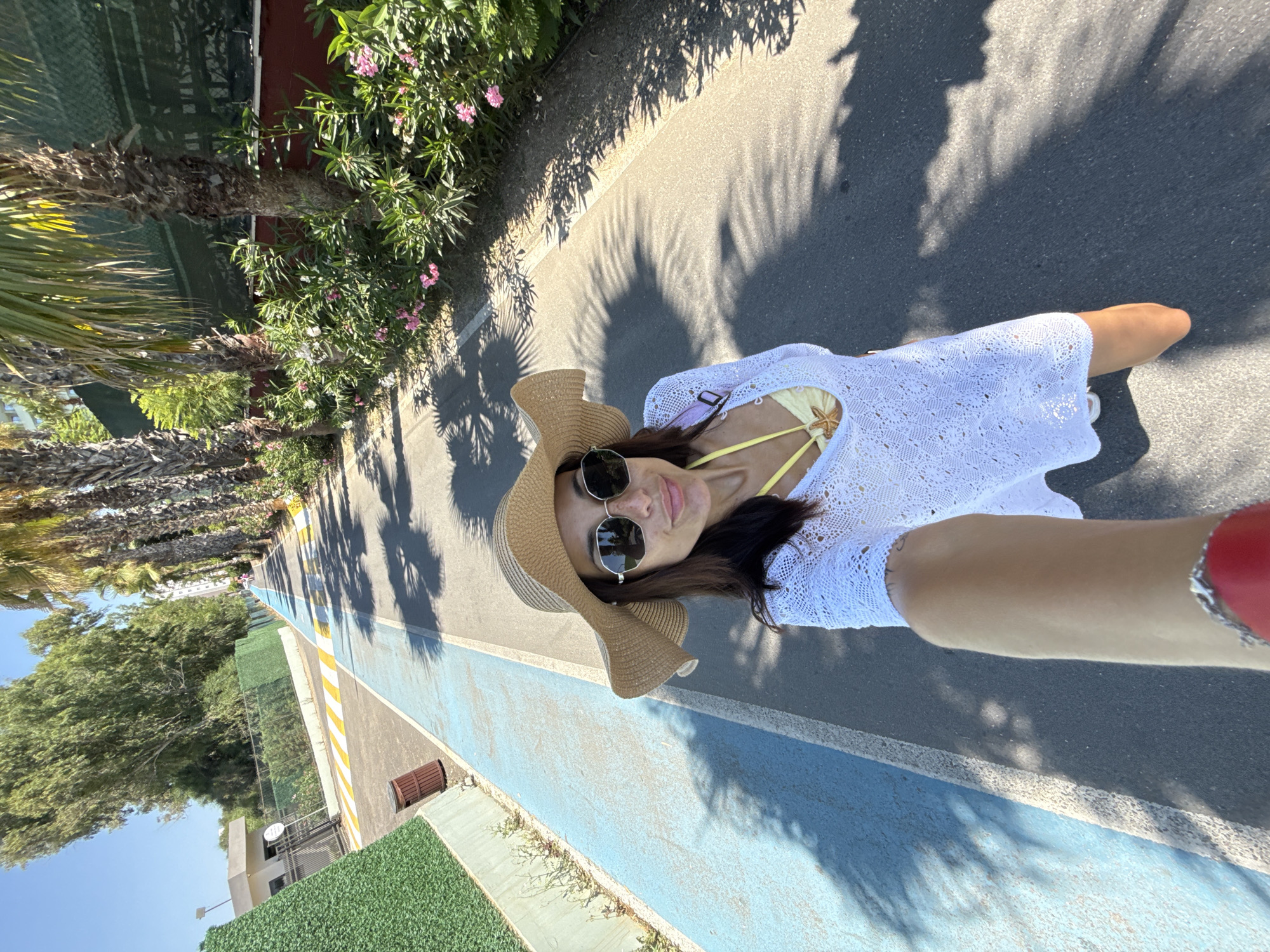 In a full-body selfie taken outdoors, a woman stands on a paved path lined with palm trees and foliage. She is wearing a wide-brimmed straw hat, sunglasses, and a white crocheted cover-up over a yellow bikini. A starfish pendant is visible on a necklace. The path is marked with a blue stripe on the left side. In the background, a green hedge runs along a fence, and a building is visible further down the path. Shadows of palm fronds cast dappled patterns on the ground. The scene appears to be in a warm, sunny climate, likely a resort area in Unknown, Türkiye.