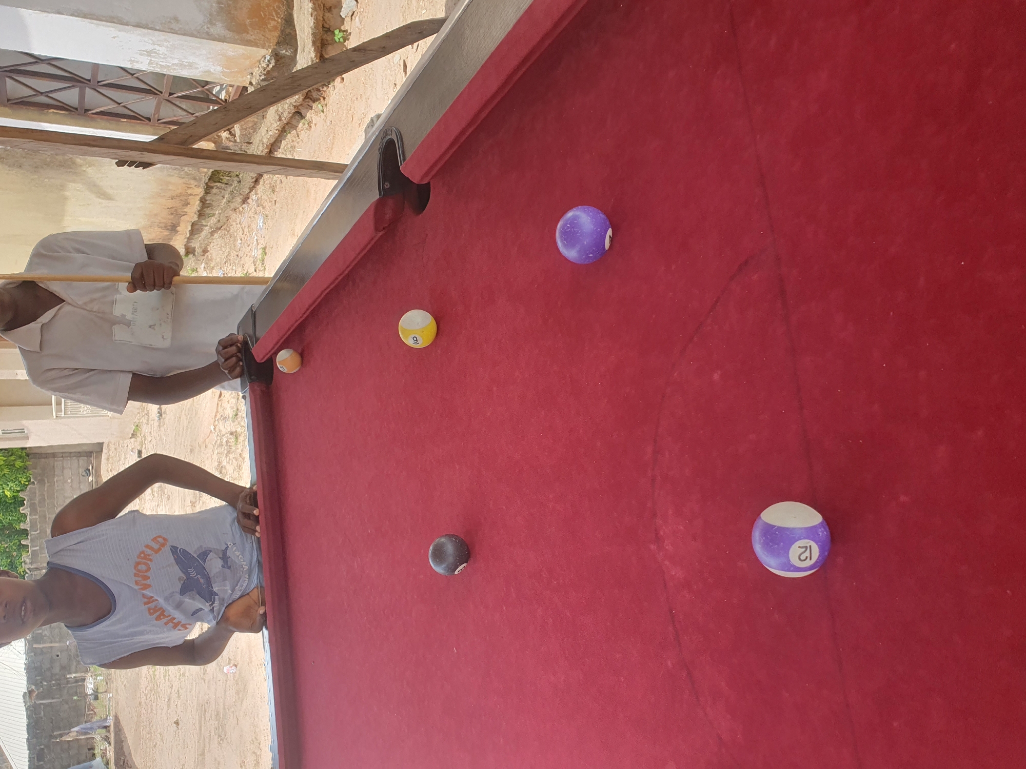 Two young males, likely teenagers, stand behind a red felt pool table outdoors. The male on the left, wearing a light blue tank top with a shark graphic and the words "SHARK WORLD," rests his hands on the table. The male on the right, in a light-colored collared shirt, holds a cue stick. Several billiard balls are scattered across the table: a black 8-ball, a yellow 9-ball, a purple and white striped 12-ball, and a solid orange ball. The background shows a dirt ground, a light-colored wall with a wooden structure and a window, and possibly a building. The overall scene suggests a casual game of pool taking place in Lokoja, Nigeria.