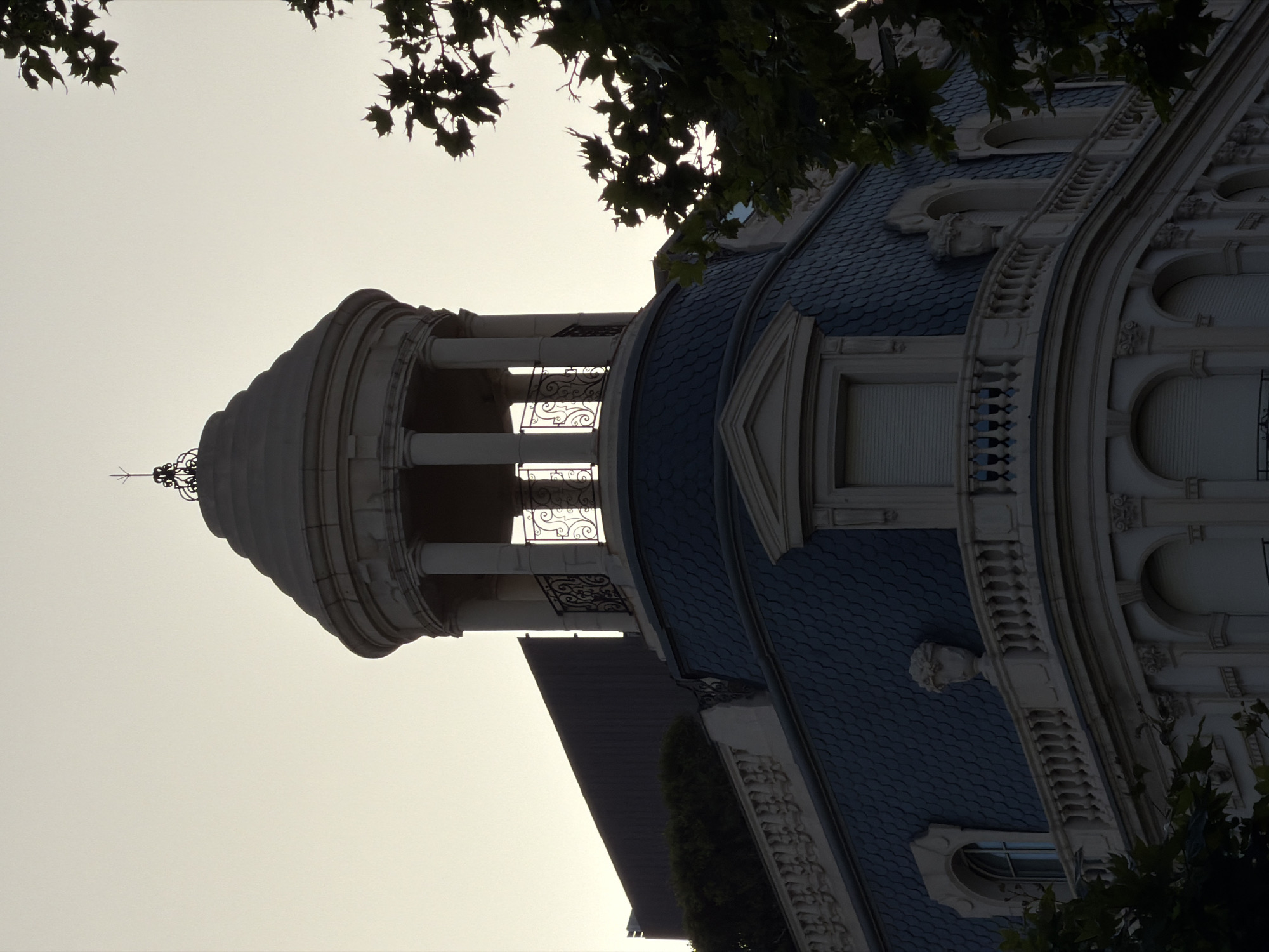 The photograph captures a section of an ornate building's rooftop and upper facade against a pale, overcast sky, likely at dusk or dawn. The architectural style appears to be French Beaux-Arts, characterized by a curved mansard roof covered in dark, overlapping shingles, a semi-circular dome-like structure with white columns and decorative railings, and finely detailed stonework. A large, dark, flat-topped structure, possibly a more modern addition or part of an adjacent building, juts out from behind the mansard roof on the left side of the image. Foliage from trees, silhouetted against the sky, frames the right side of the image, obscuring some of the building's details. The overall impression is one of grandeur and historical architecture. This specific building is located on Passeig de Gràcia in Barcelona, Spain, a renowned avenue known for its modernist architecture and luxury shops.